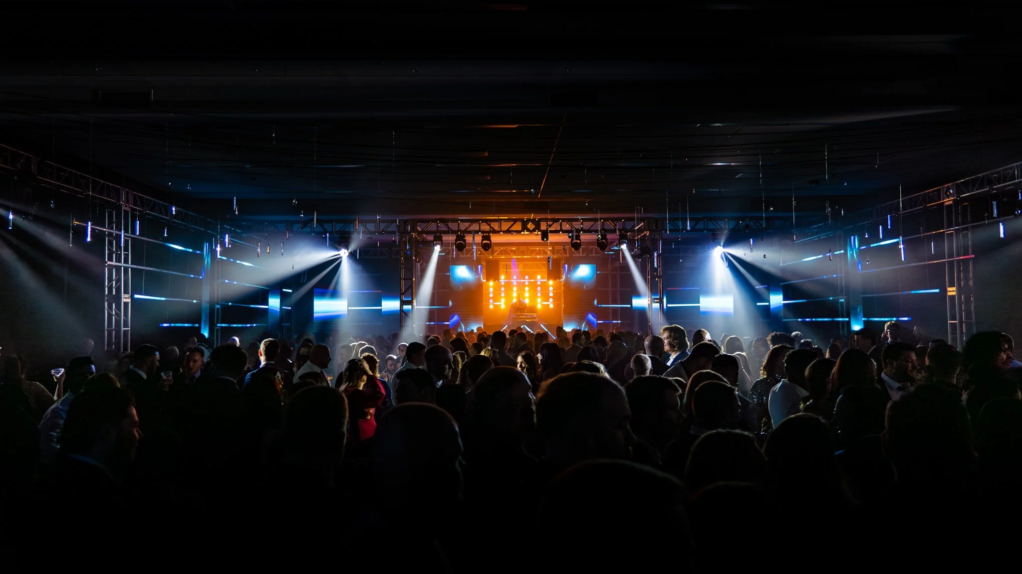 Crowd of people dancing and socializing at a concert or nightclub with stage lighting and DJ booth in the background.