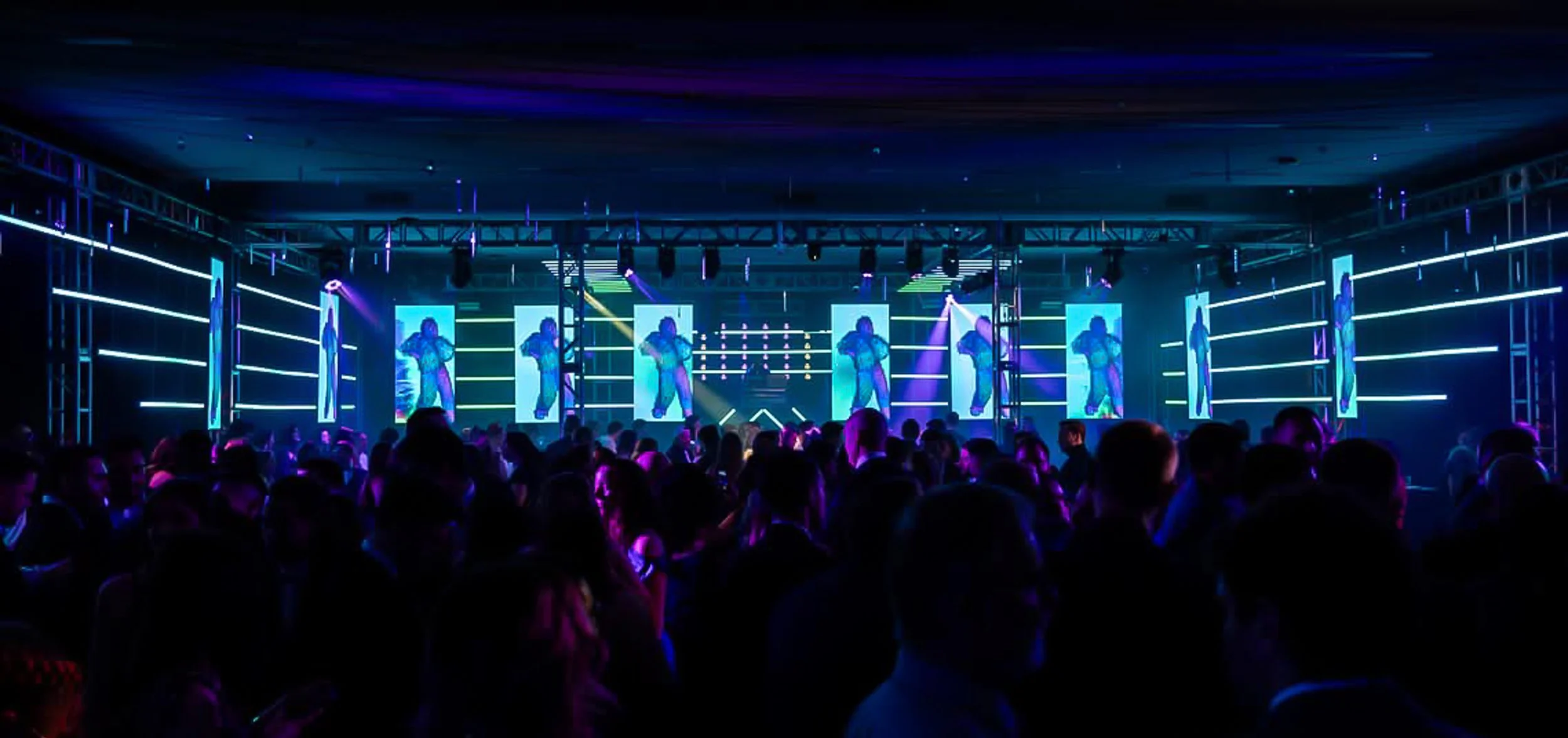 A crowded dance floor with people watching a stage with multiple large screens showing a dancer, neon lights, and colorful laser lighting effects in a nightclub or concert venue.