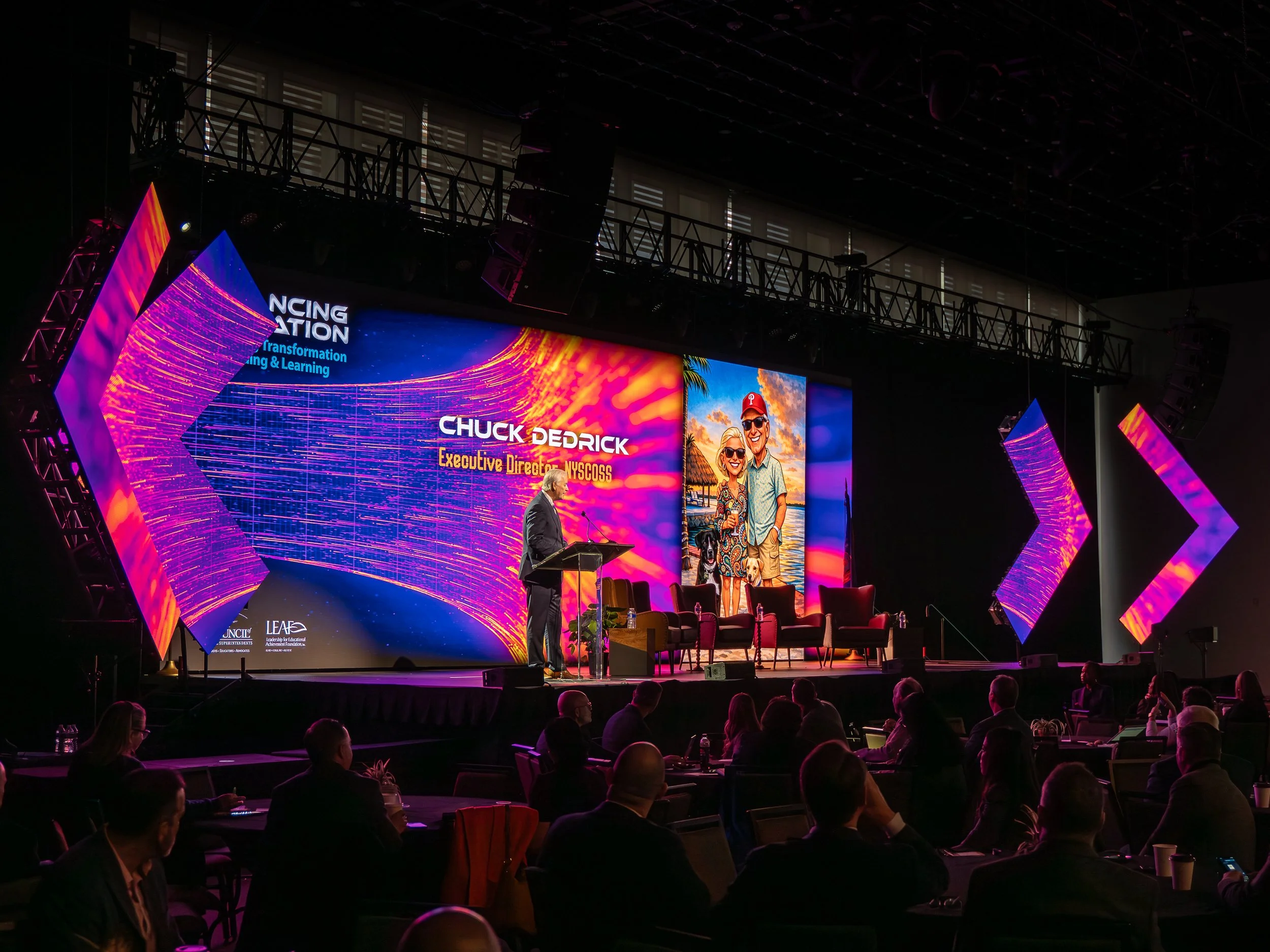A man standing at a podium on stage giving a presentation at a conference. The stage background displays the name Chuck DeDrick and his title, with a large colorful digital screen showing graphics and an image of a couple on vacation. Attendees sit a