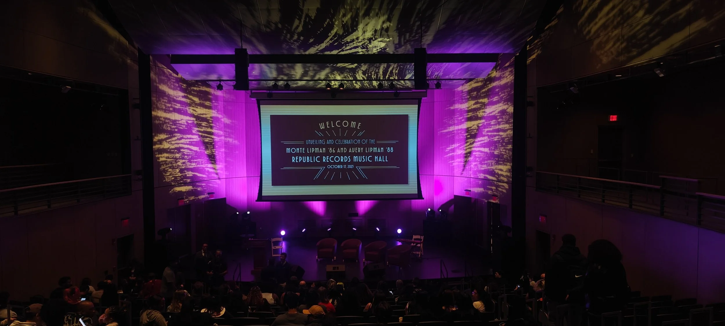 The stage of a music hall with purple lighting, a large screen displaying a welcome message for the unveiling and celebration of Monte Lipman '86 and Aubrey Lipman '88 at the Republic Records Music Hall on October 17, 2023, and some chairs and people