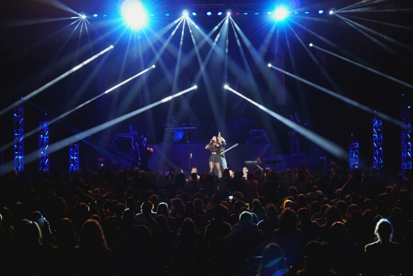 A live concert with performers on stage, bright blue and white stage lights, and an audience watching.