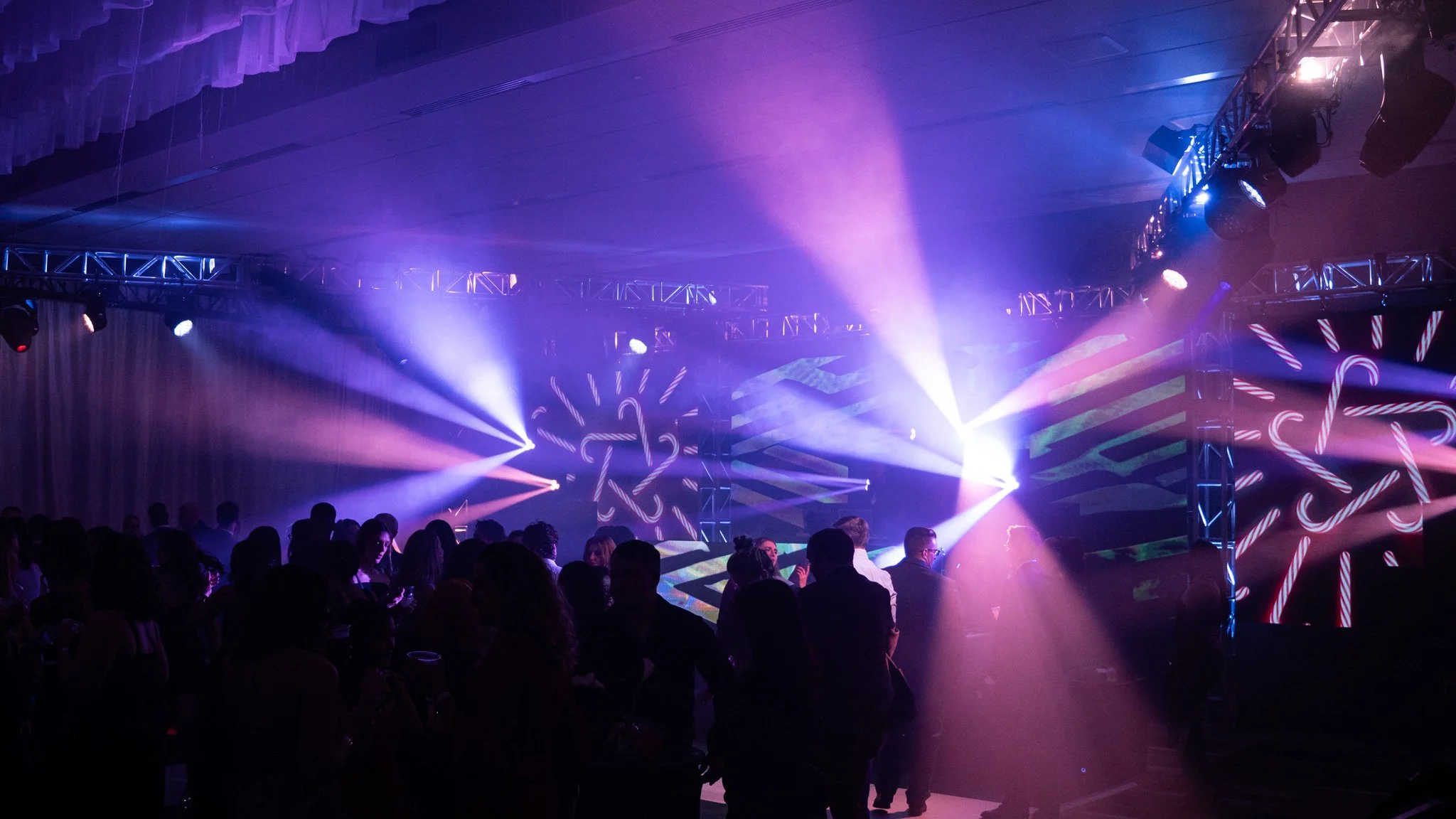 A crowd of people dancing and socializing at a party with colorful lights and a large LED display in the background.