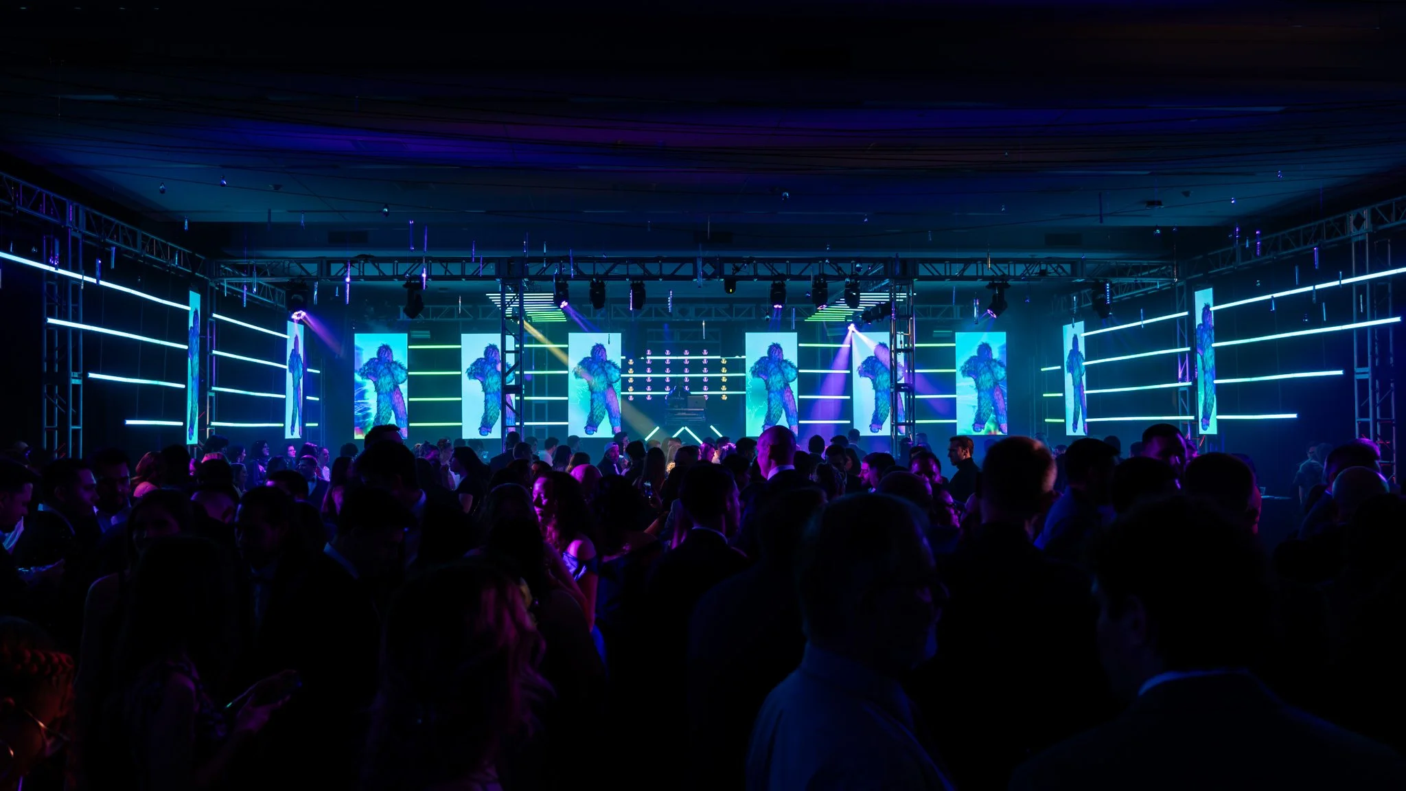 A busy nightclub with a DJ on stage and large screen displays, illuminated by colorful neon lights, surrounded by a crowd of people dancing and socializing.