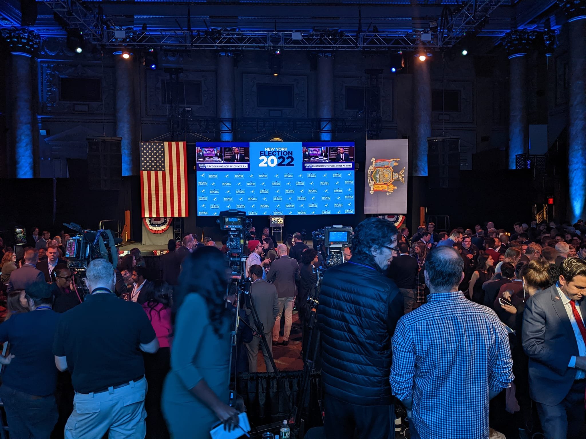Crowd of people at a news conference or election event with a large screen displaying 'New York Election 2022' and American flags in the background.
