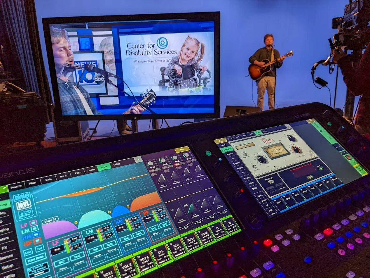 A live music performance in a studio with a large screen showing a child in a wheelchair and the logo of the Center for Disability Services, with two musicians playing guitar and a camera operator recording. In the foreground, a digital mixing consol