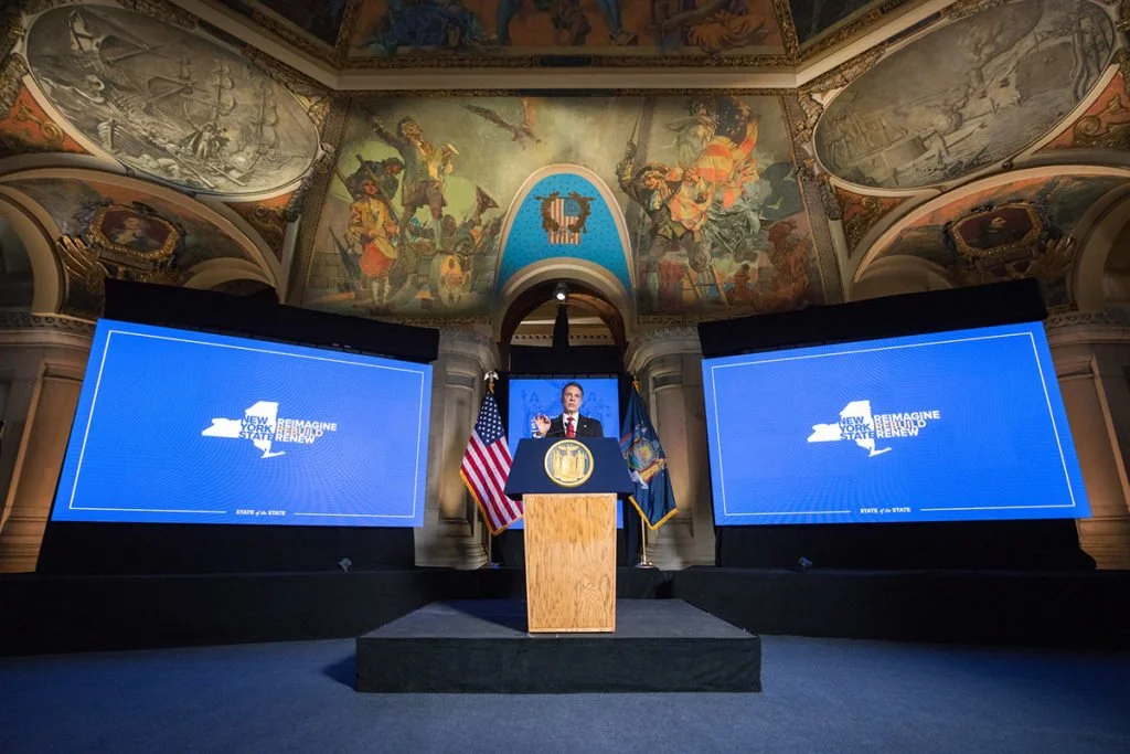 A man at a podium speaking at an indoor event with two large screens displaying the slogan 'New York State ReImagine, Renew, Thrive.' The backdrop features American and state flags, and a decorative stained glass ceiling is visible above.