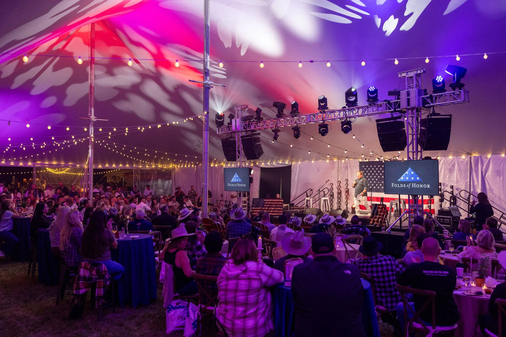People attending an indoor concert or event in a large tent with string lights and colorful lighting, a stage with a speaker speaking into a microphone, and a large American flag backdrop.