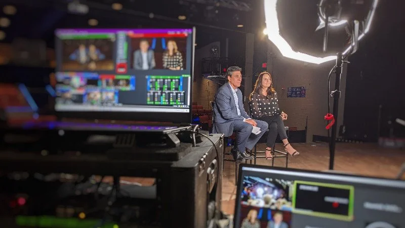 Two people sitting on a stage during a video recording or interview, with professional lighting and camera equipment visible.