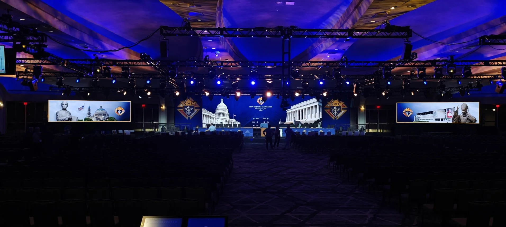 Conference hall with a stage, large screens showing images of the U.S. Capitol, a statue, and a building, decorated with the Knights of Columbus symbols, features blue lighting and a few attendees setting up.