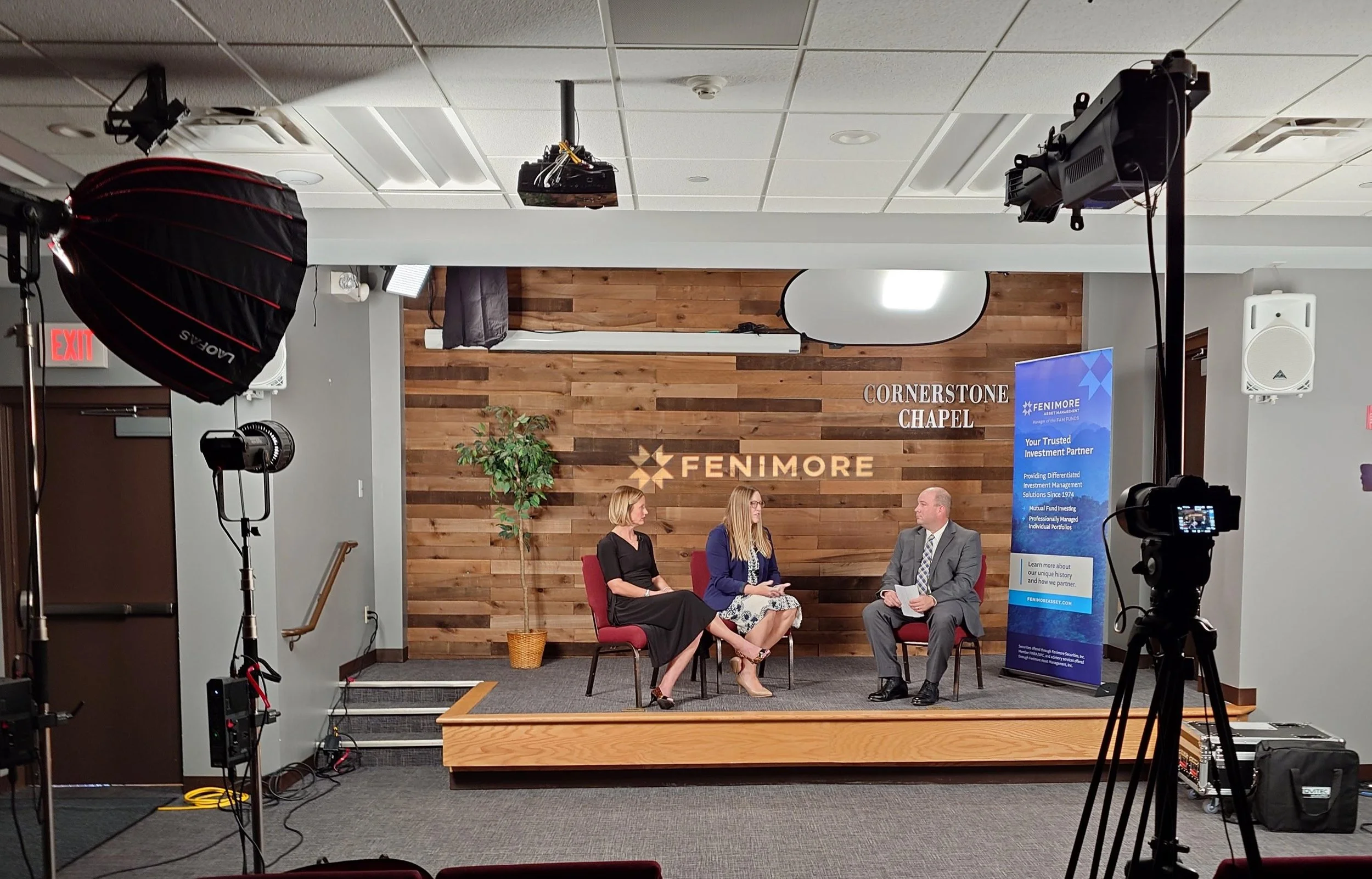 A panel discussion taking place in a room with wooden wall panels, where three people are seated on stage. Two women and one man are engaged in conversation, with cameras and lighting equipment set up around them. A sign reads 'CORNERSTONE CHAPEL,' a