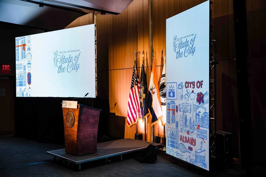 A stage with a wooden podium, a large screen displaying 'State of the City' for Albany, with illustrations and flags of the United States, New York state, and Albany city in the background.