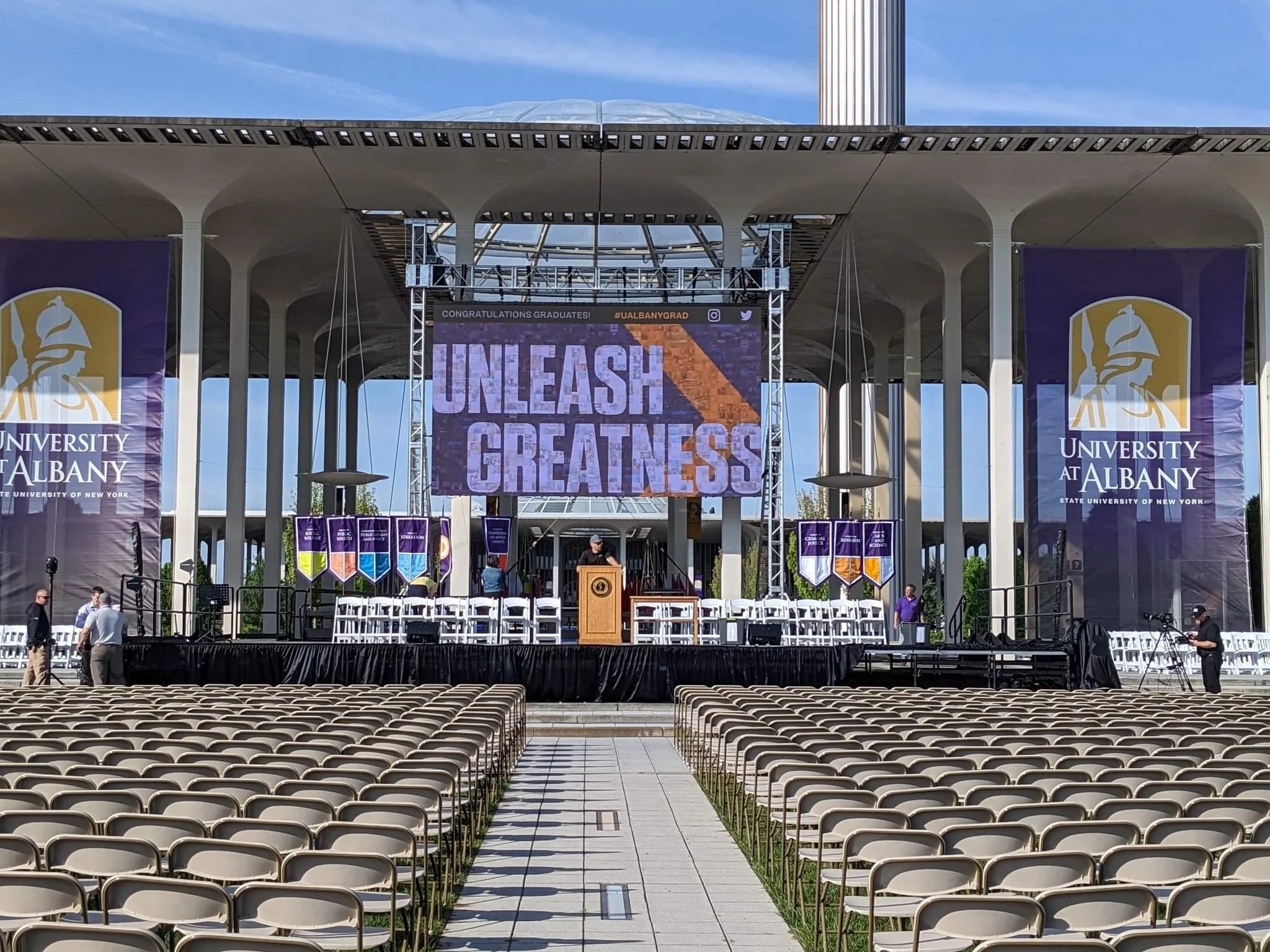 Outdoor graduation stage at University at Albany with large banners and a screen saying 'Unleash Greatness'.