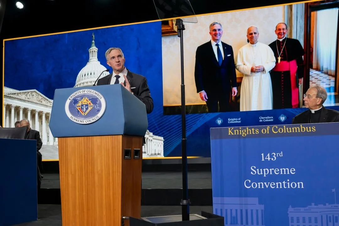 A man speaking at a podium with the Knights of Columbus Supreme Council logo, with an enlarged screen behind him showing four men, including a religious figure in white robes, a man in a black suit, and a bishop in black and red clerical attire, duri