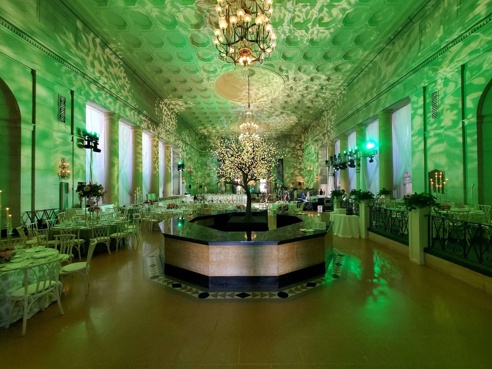 Elegant banquet hall decorated with green lighting, a central illuminated tree, round tables with white chairs, and chandeliers.