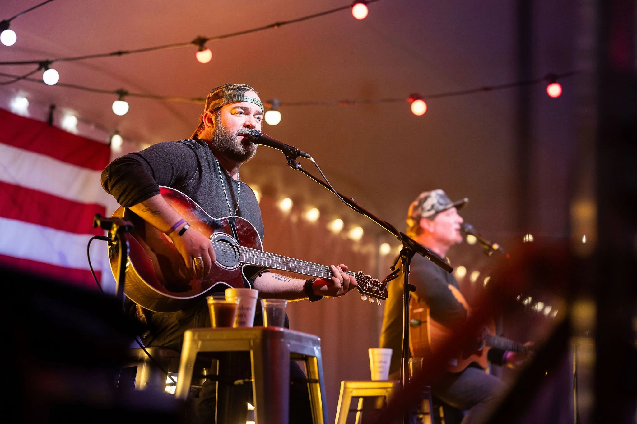 Two musicians playing acoustic guitars and singing into microphones on stage at a nighttime event, with string lights overhead and a large American flag in the background.