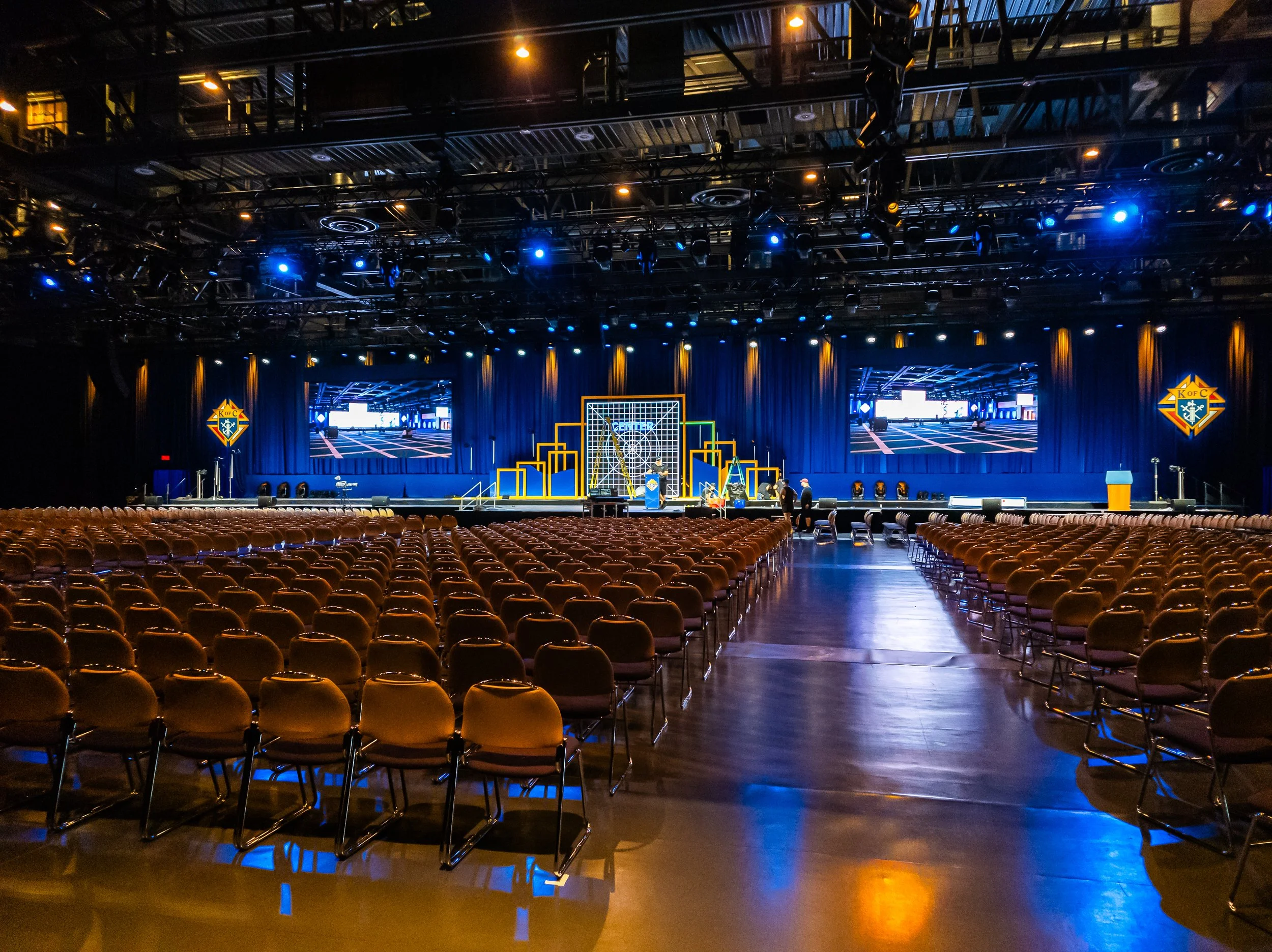 An empty conference hall with rows of chairs facing a stage, which has large screens, a podium, and decorative geometric structures; the stage area features a backdrop with the KOF C logo and is illuminated with blue and yellow lighting.