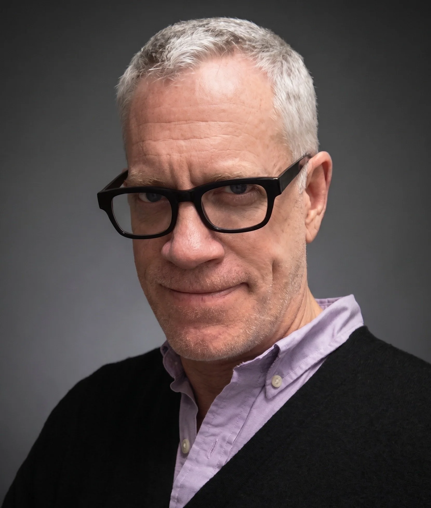 Close-up of a smiling older man with short gray hair and black glasses, wearing a purple shirt and dark sweater, against a dark gray background.