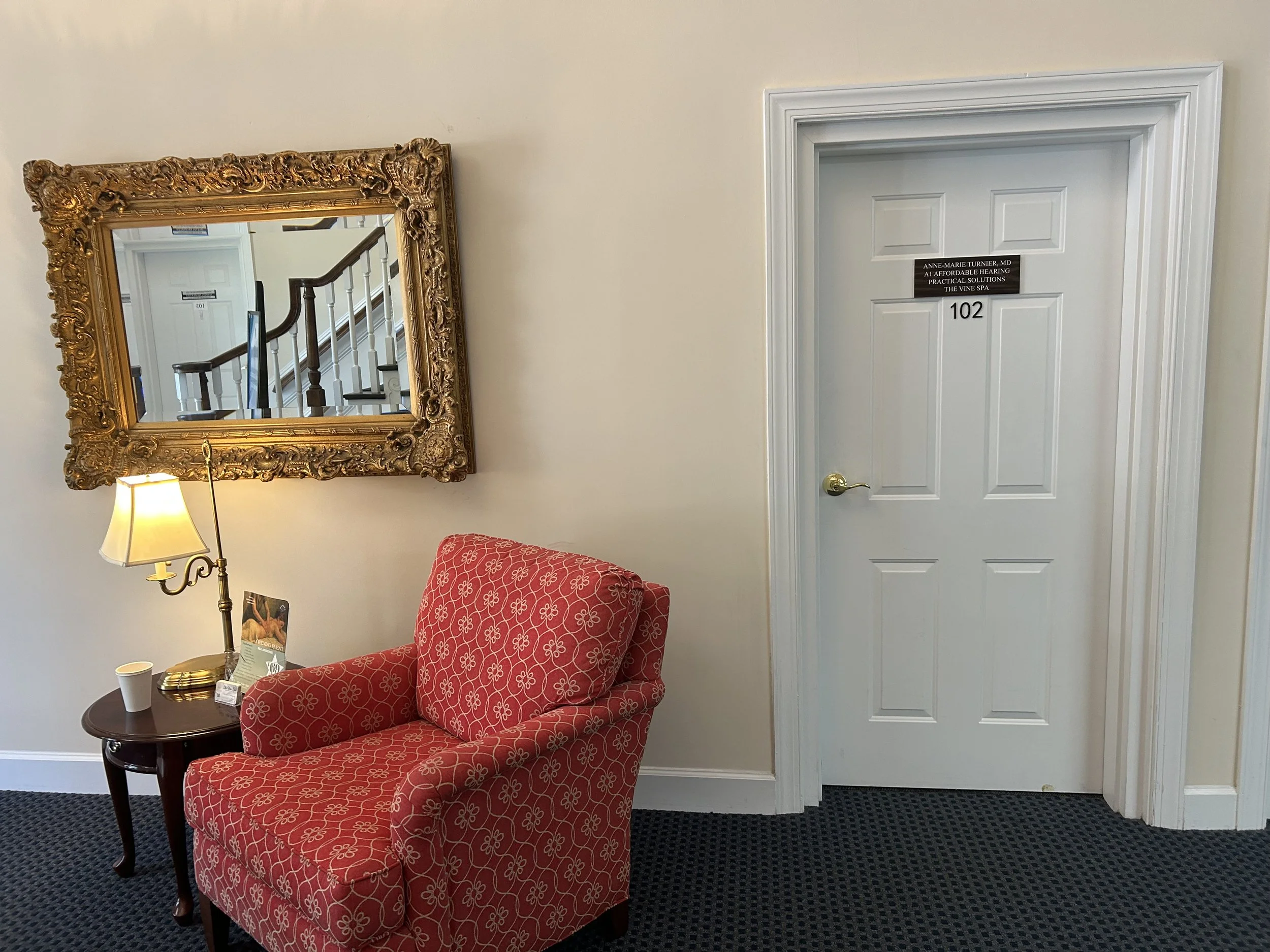 A waiting area with a red patterned armchair, a small wooden side table with a lamp, a paper cup, and framed promotional material, a large ornate gold-framed mirror on the wall, and a closed white door with a black sign and room number '102'.