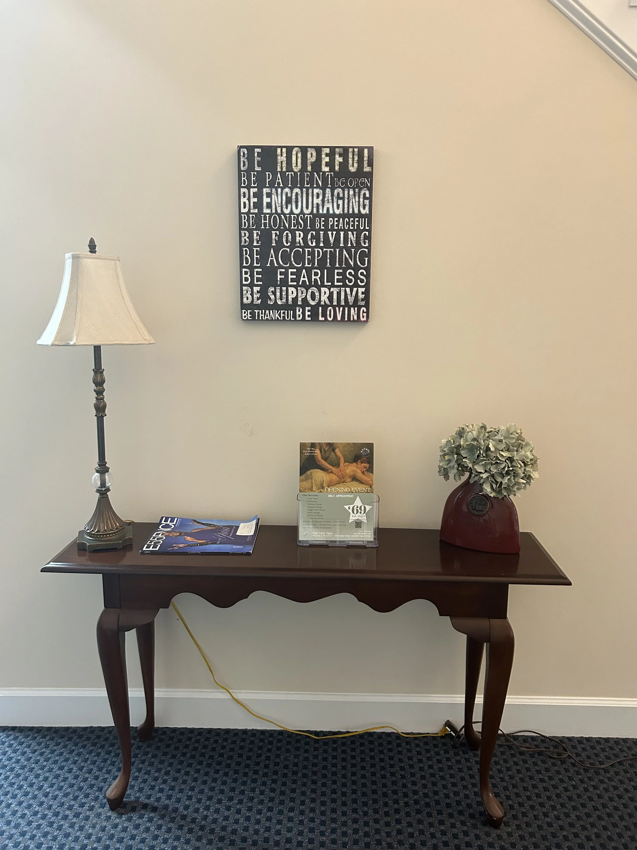 Wooden table with a lamp, a magazine, a promotional sign, and a flower vase with white flowers against a beige wall with motivational quote artwork.