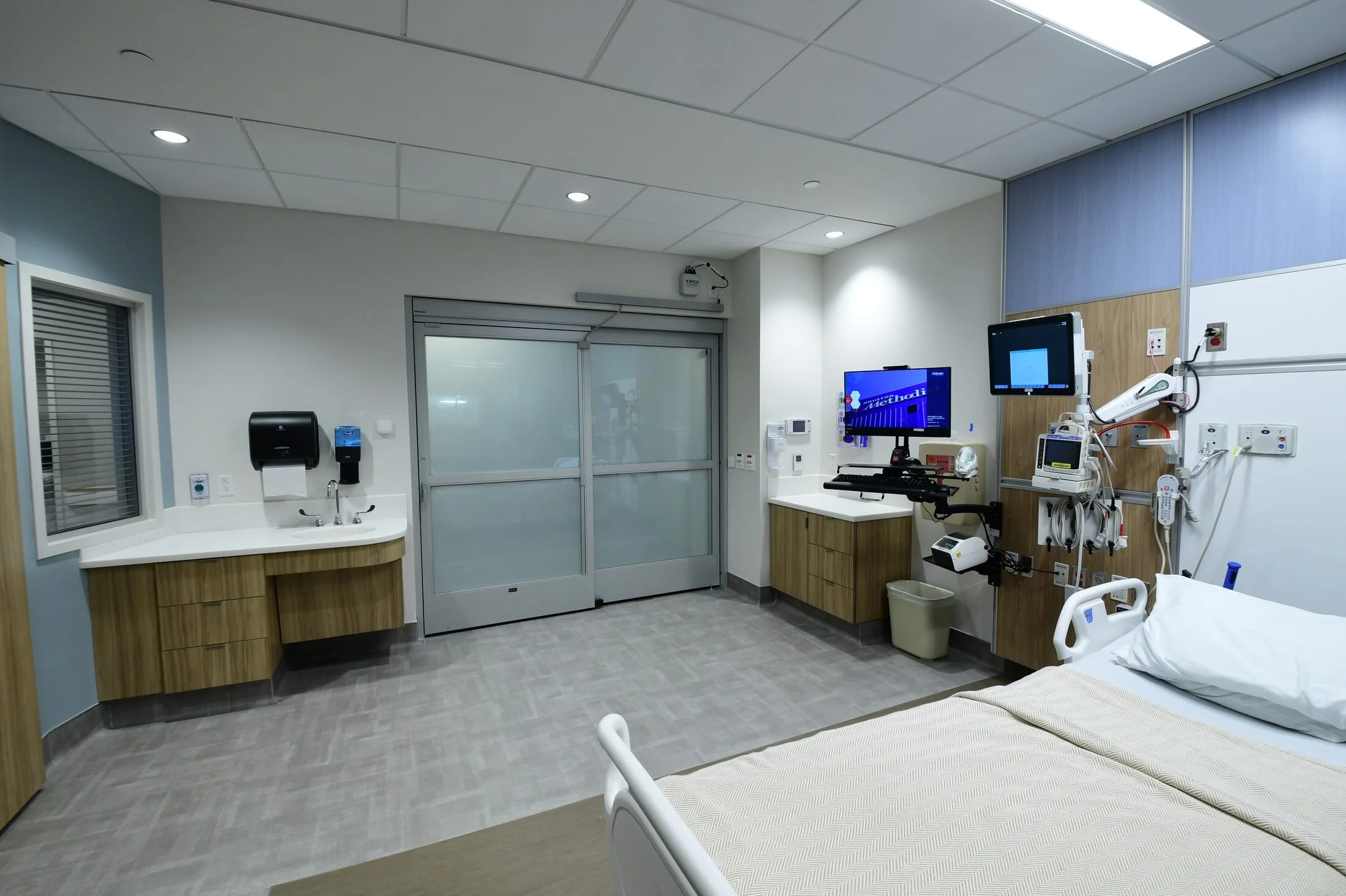 Intensive care patient room at Houston Methodist Cypress, seen from across the bed, with a SmartLayer switchable glass door in frosted privacy mode at the corridor face.