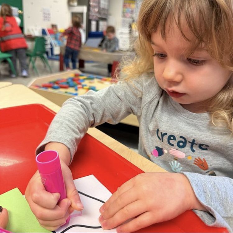 A young girl with long curly blond hair drawing on a piece of paper with a pink marker in a classroom or playroom. In the background, other children are engaged in activities at various tables and shelves with educational materials.