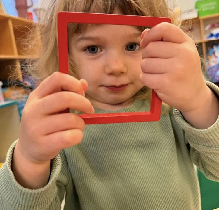 A young girl with blonde hair and blue eyes holding a red picture frame in front of her face, looking through it in a colorful room.