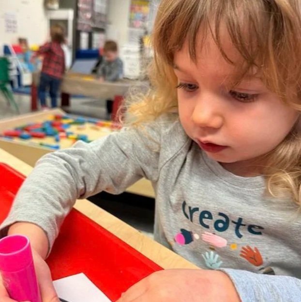 A young girl with curly blonde hair drawing with a pink marker at a table in a classroom or play area.