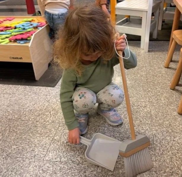 A young girl with red hair, wearing a green long-sleeve shirt and floral pants, playing with a toy broom and dustpan on a tile floor in a room with shelves and chairs.
