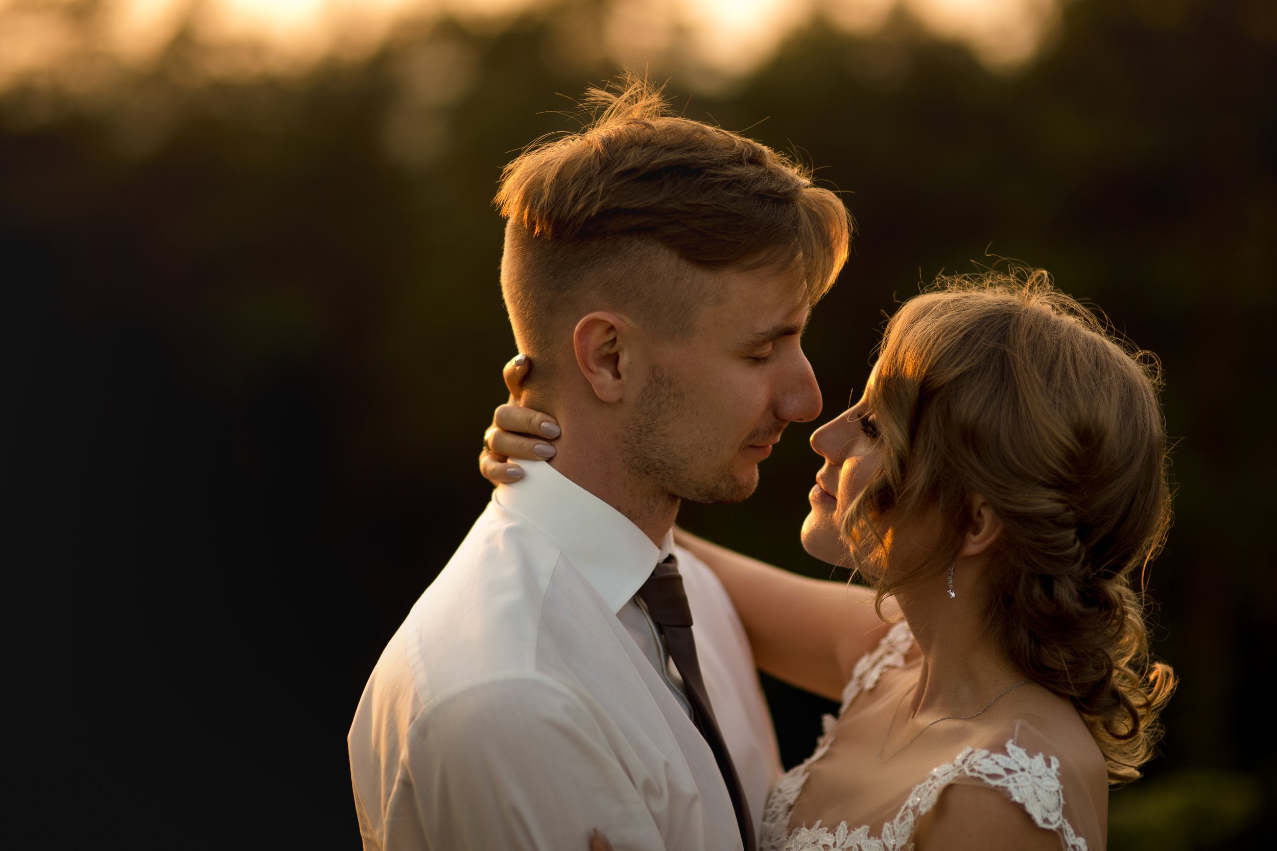 A man and woman are close together outdoors at sunset, with their faces touching. The man is wearing a white shirt and dark tie, and the woman is in a lace dress. They are embracing tenderly.