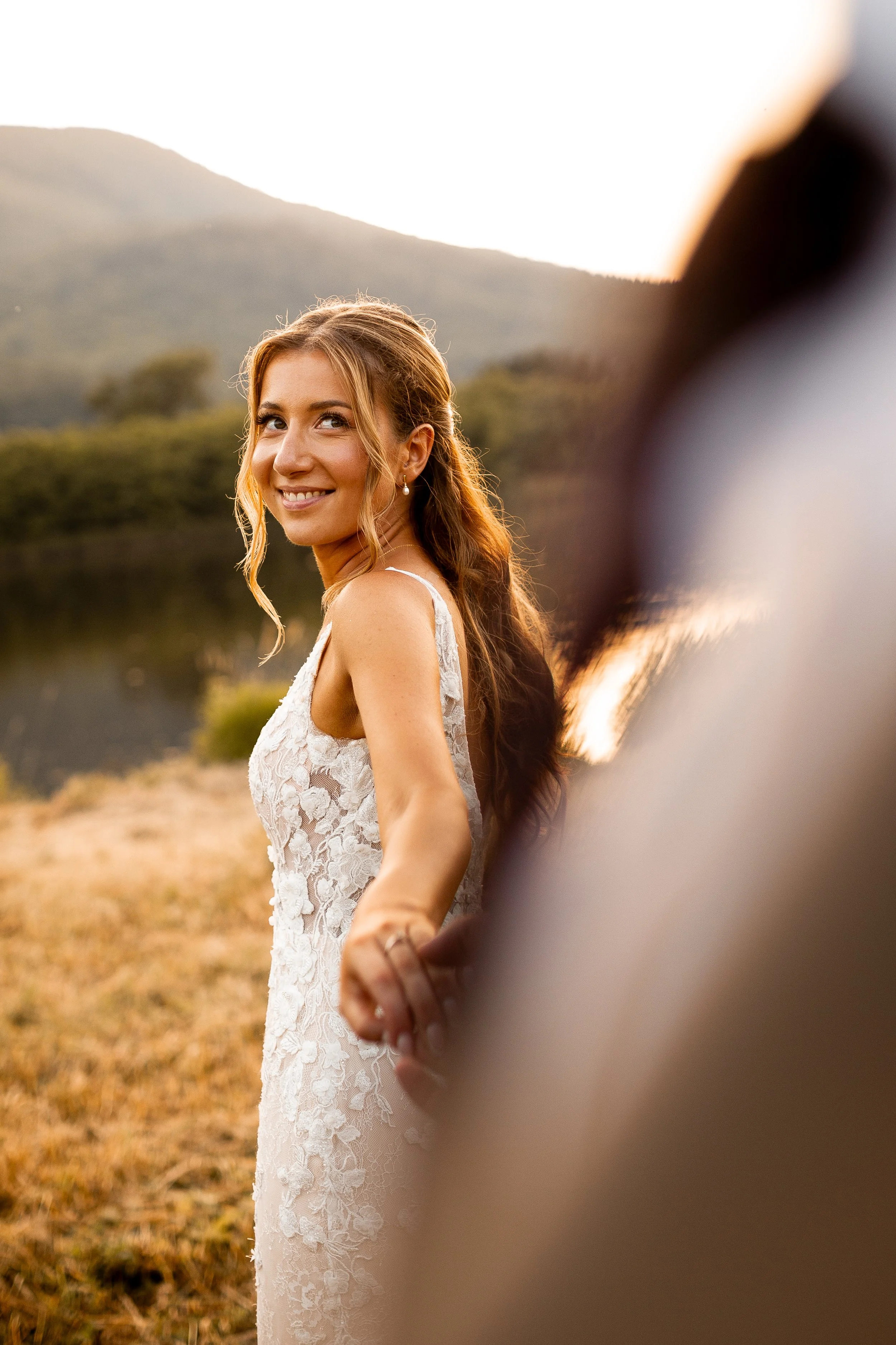 A smiling woman in a white lace dress holding hands with someone outside near a lake and mountains at sunset.