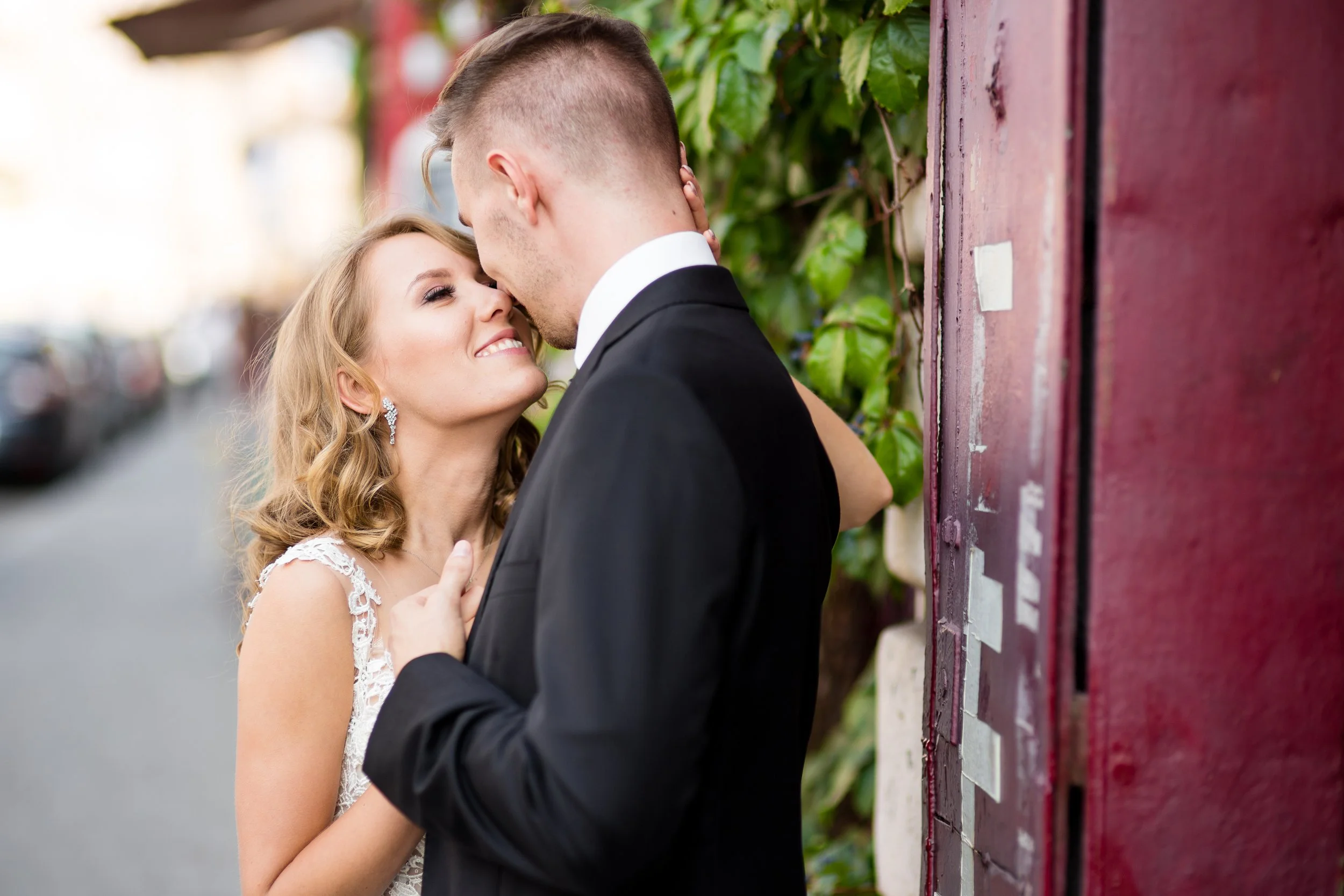 A couple dressed in wedding attire sharing a kiss outdoors, with the woman wearing a lace dress and the man in a black suit, standing next to a red wall with green foliage.