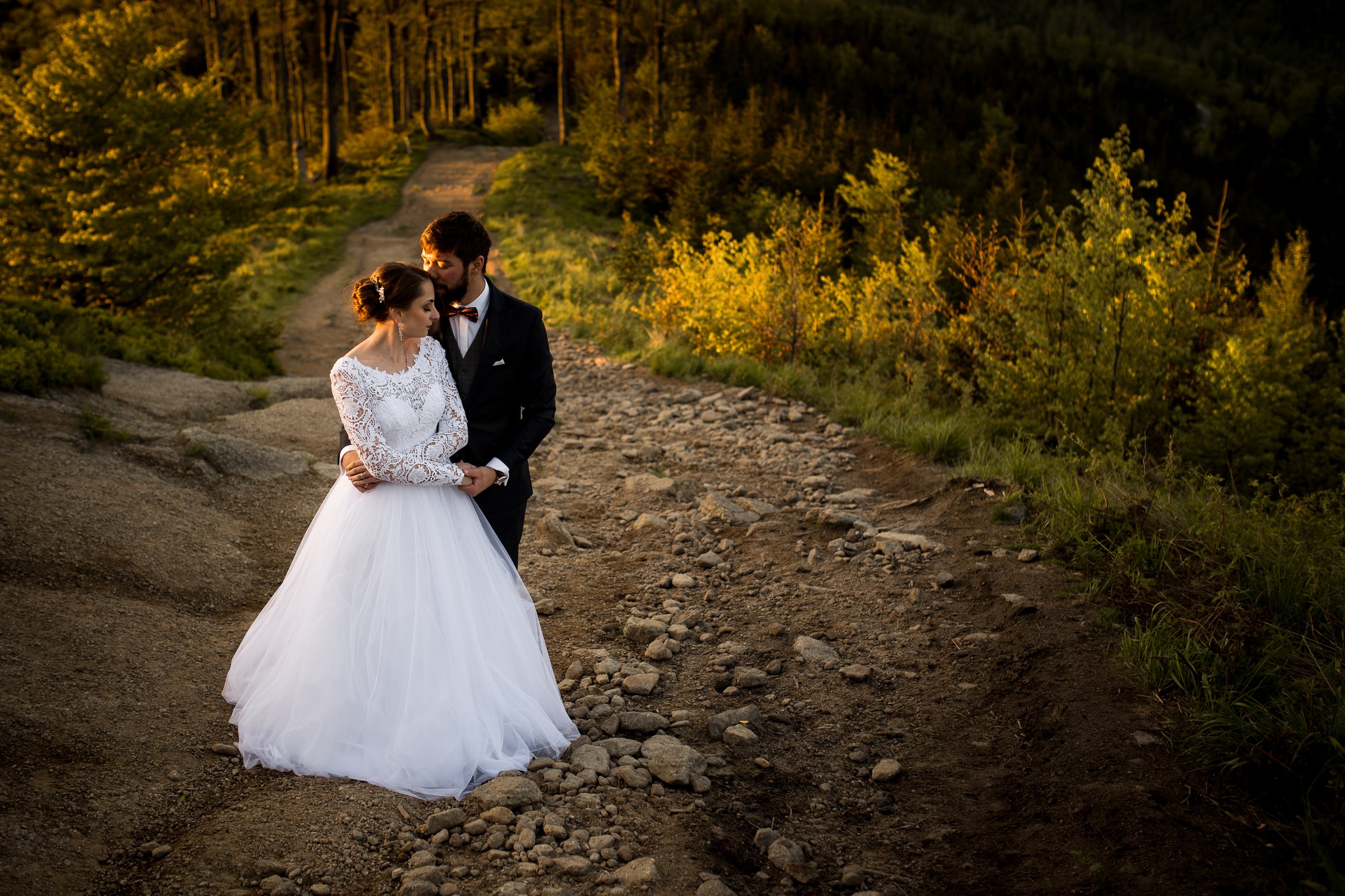 A bride and groom standing together on a rocky trail in a forested area during sunset, gazing at each other.
