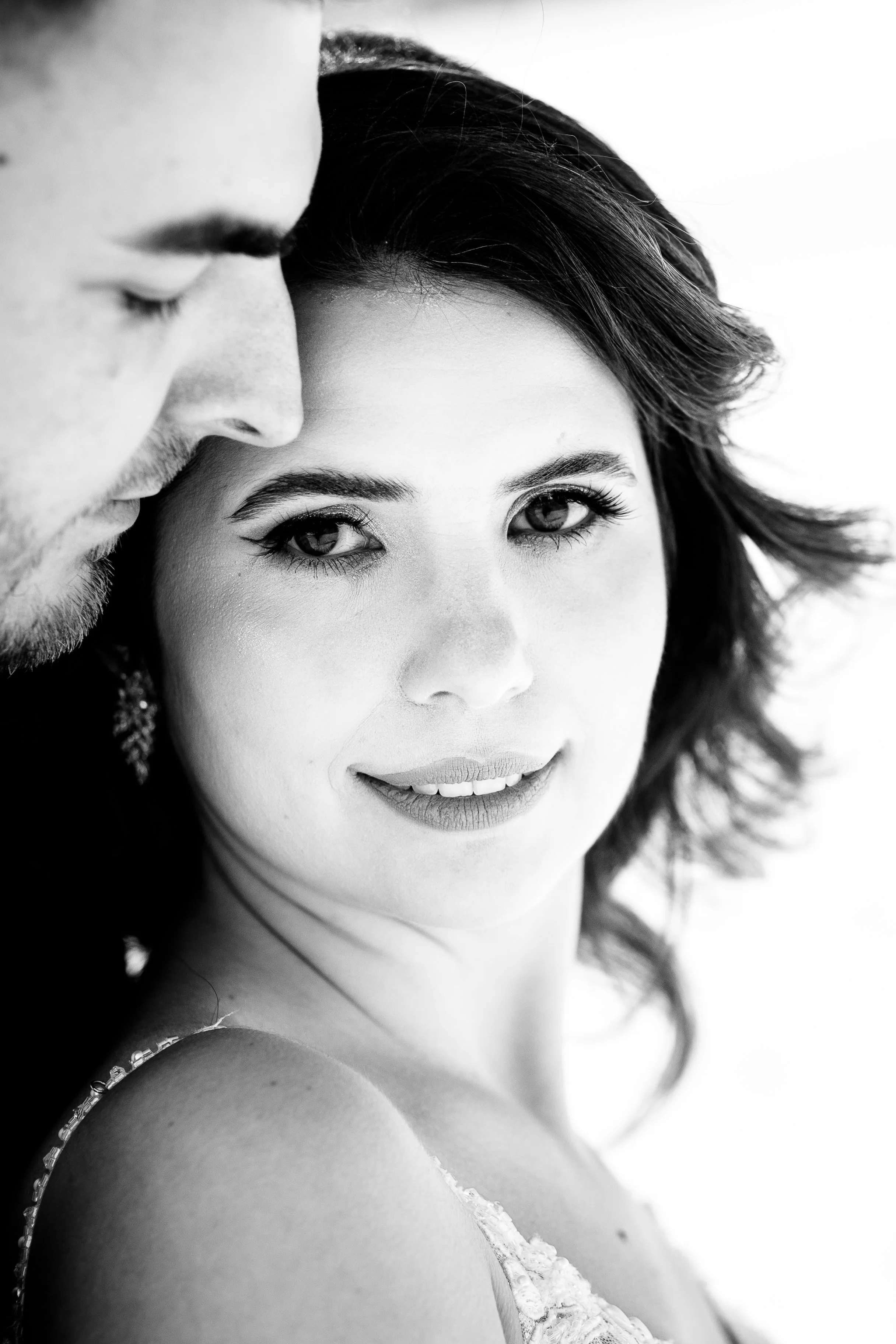 A black and white close-up portrait of a woman with dark hair and makeup, looking directly at the camera, with a man partially visible beside her.