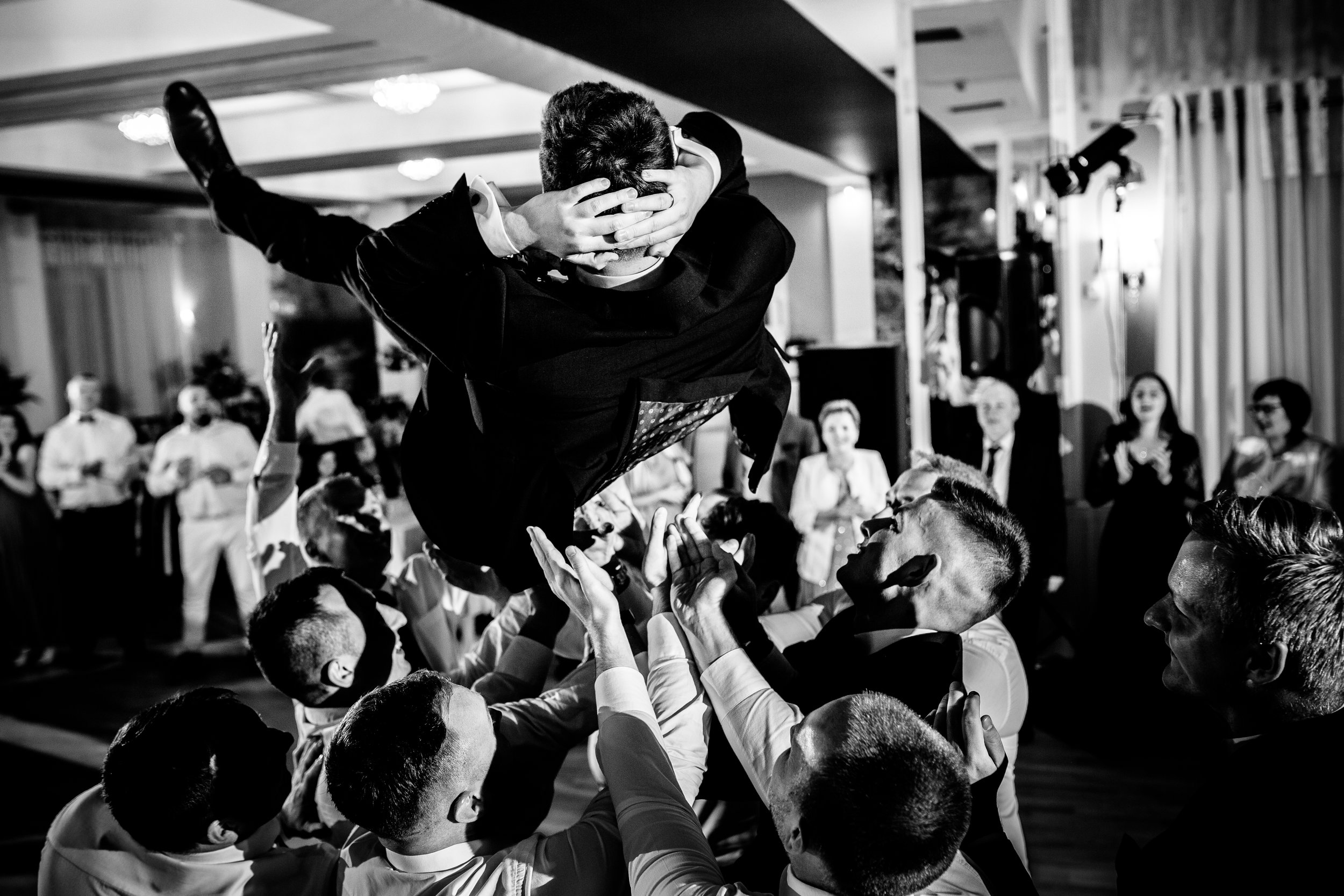 A man is being lifted into the air by others during a celebration, with the man holding his hands to his head and crowd clapping and watching in the background.