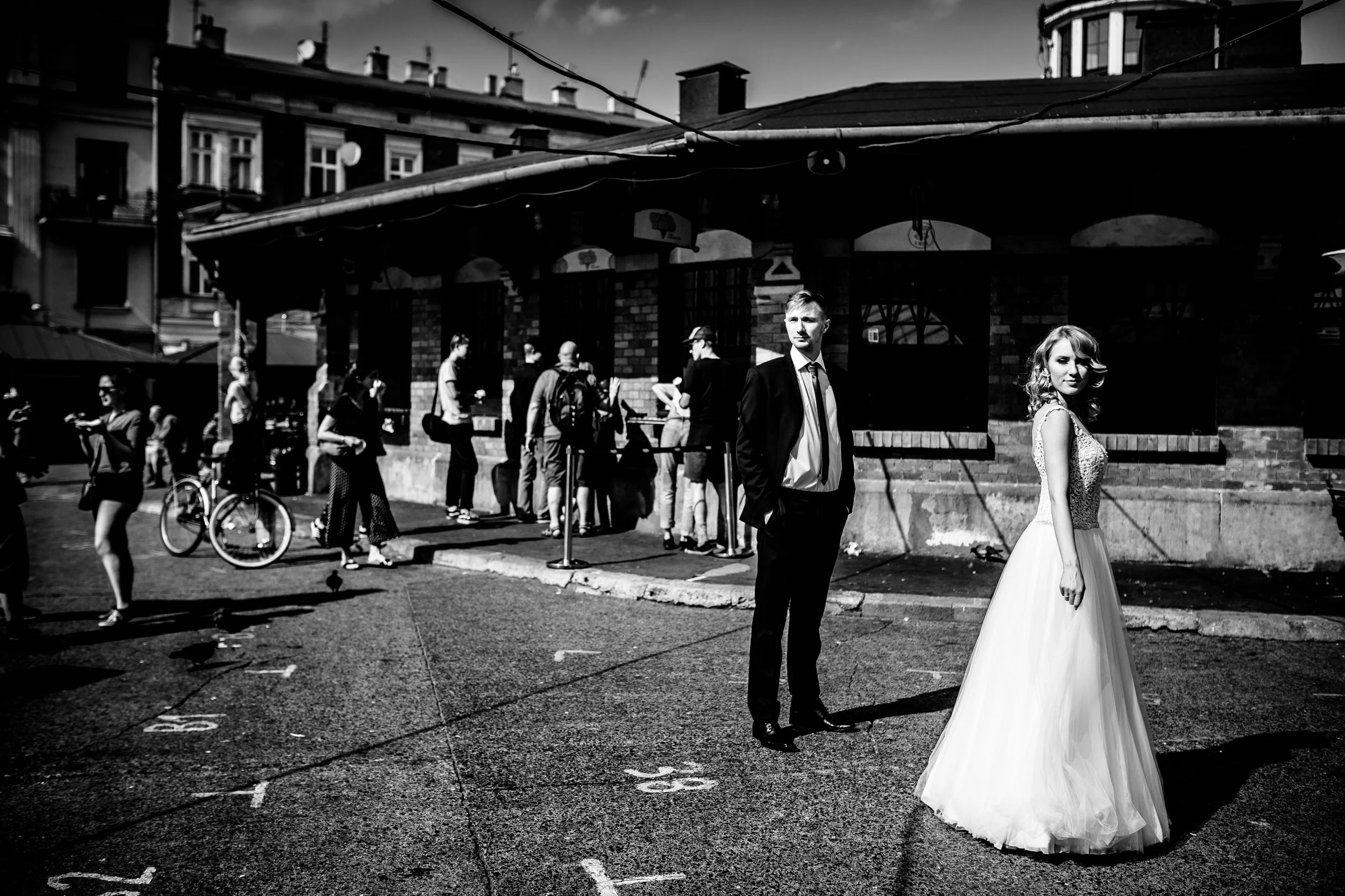 In a black and white photo, a woman in a wedding dress and a man in a suit stand in a parking lot with numbered spaces. The man is in the foreground looking away, while the woman is slightly behind him, facing the camera. In the background, people ar