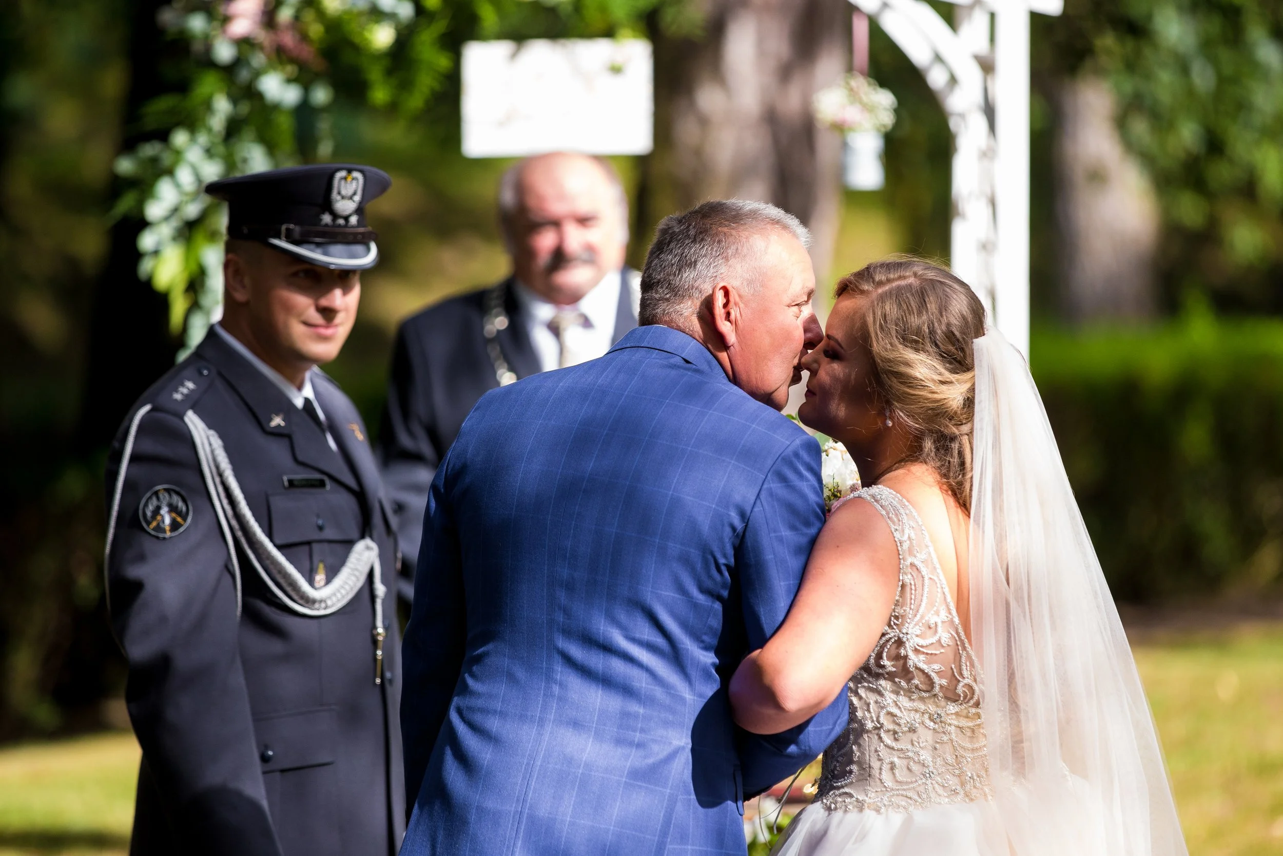 A bride and a man, possibly her father, share an intimate moment during a wedding ceremony outdoors, with a military officer and an officiant observing in the background.
