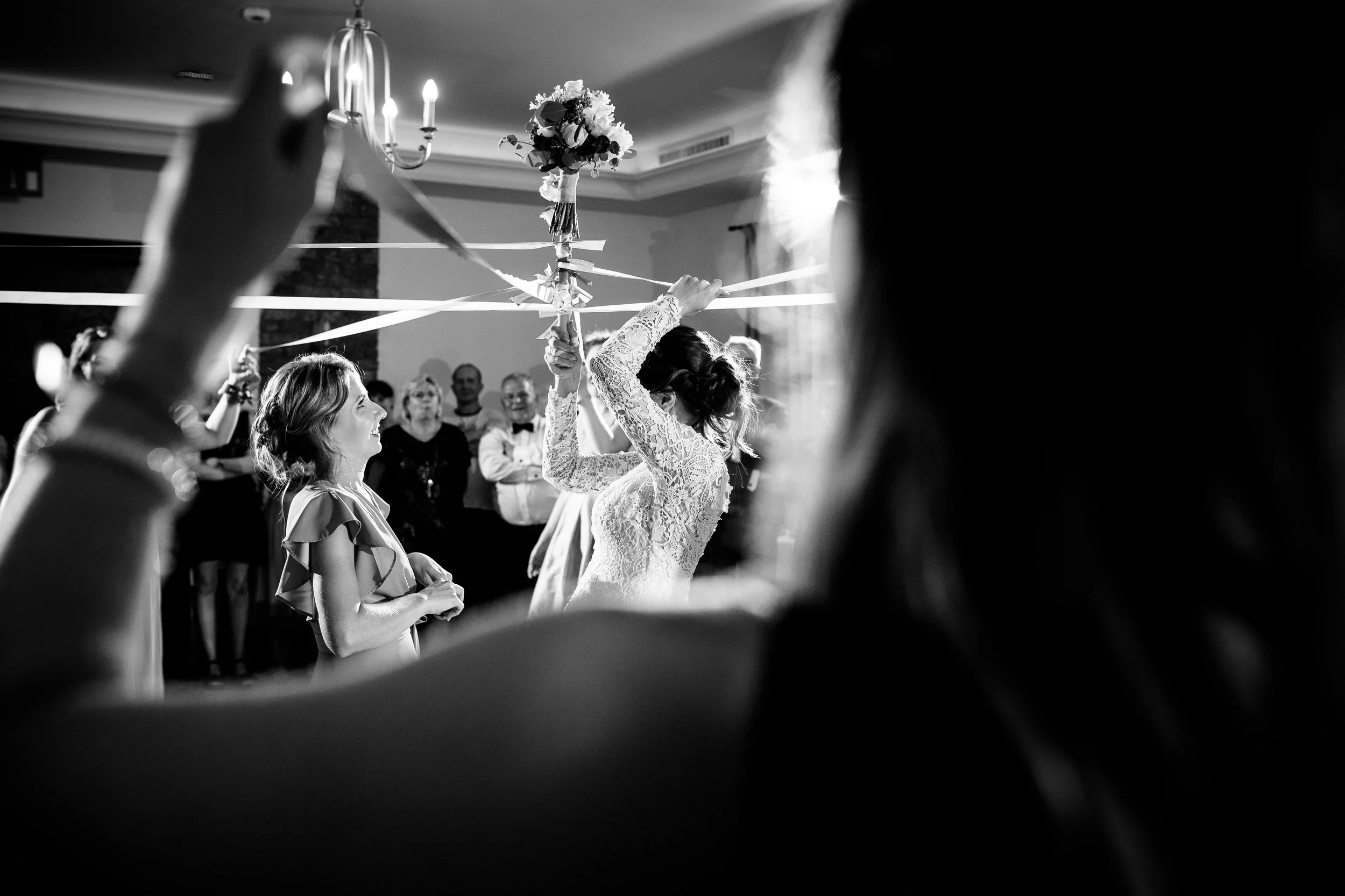 A bride and guests celebrate during a wedding reception, with a bouquet toss in progress and many guests watching, in a well-lit room.