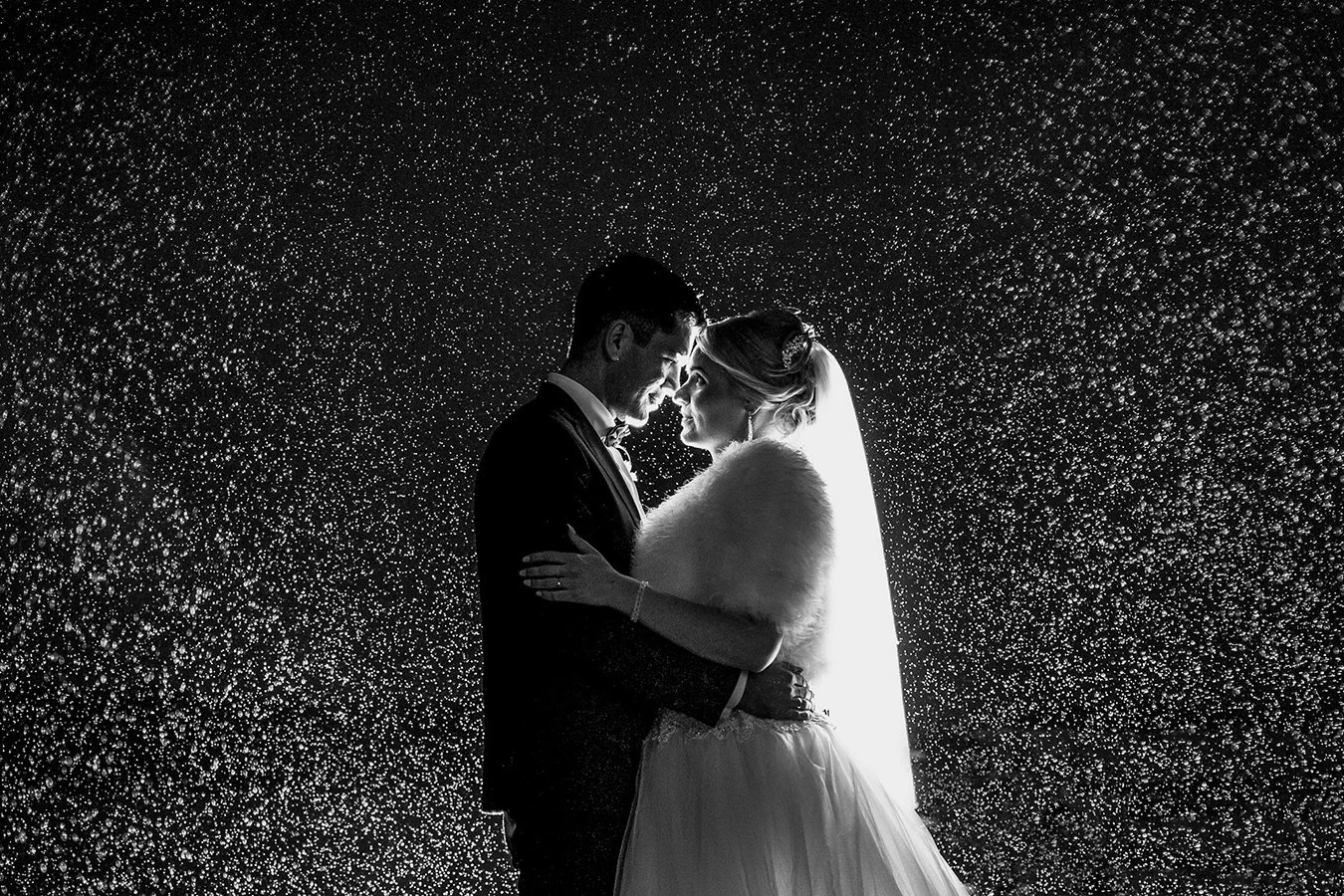 Czarno-białe zdjęcie przedstawiające pannę młodą i pana młodego stojących blisko siebie, dotykających się czołami, na tle padającego deszczu, oświetlonego światłem, co tworzy dramatyczny efekt.