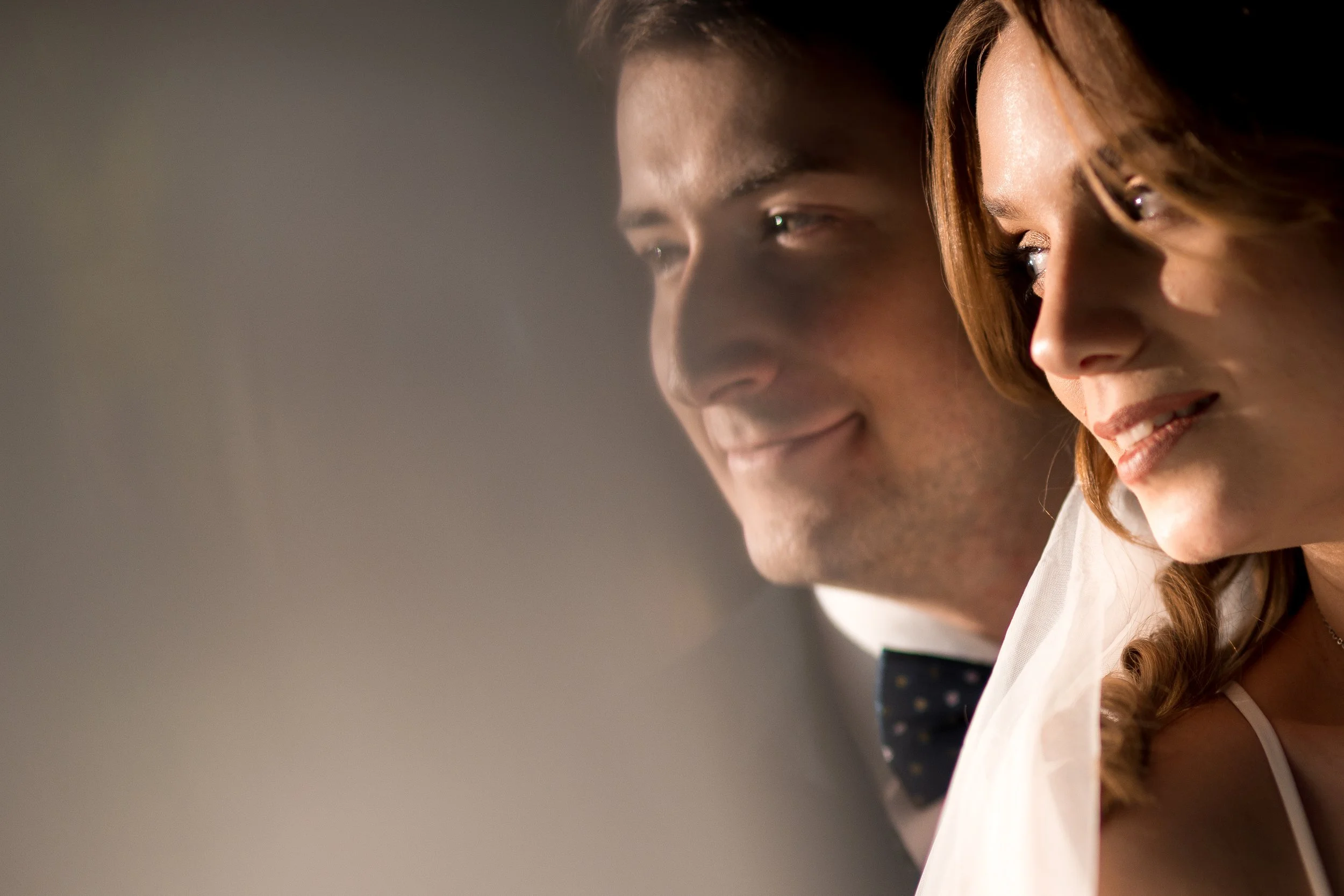 Close-up of a smiling man and woman looking to the right, with soft lighting highlighting their faces, suggesting a romantic or intimate moment.