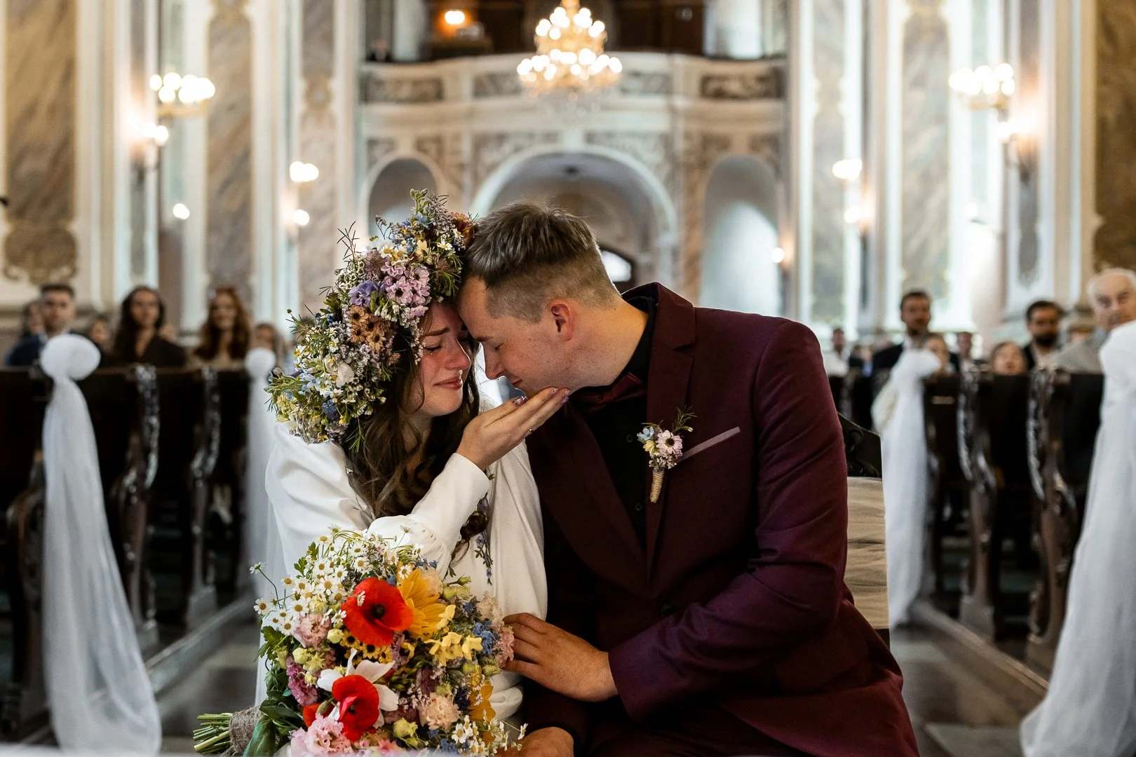 Panna młoda w wianku z kwiatów i bukiecie oraz pan młody w bordowym garniturze przeżywają wzruszającą chwilę, stykając się czołami w udekorowanym kościele podczas ceremonii ślubnej.