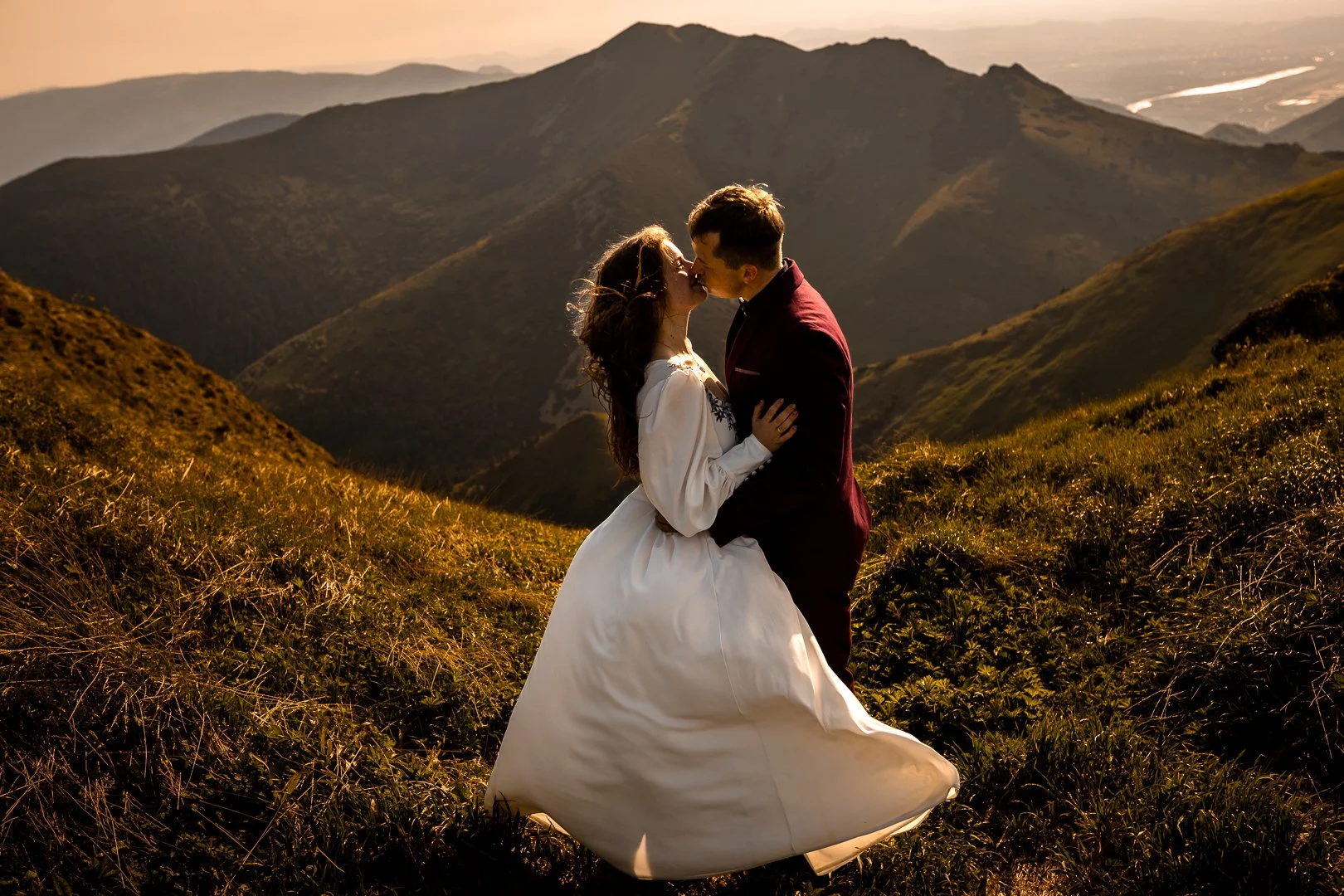 Para młoda całuje się na trawiastym zboczu wzgórza, w tle widać pasma górskie, podczas zachodu słońca.