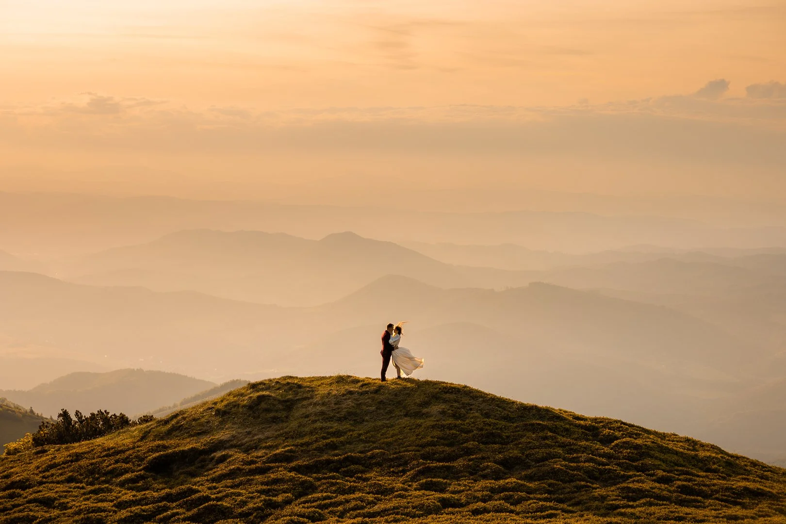 Para ubrana w strój ślubny, obejmująca się na szczycie wzgórza o zachodzie słońca, z pasmami górskimi w tle.