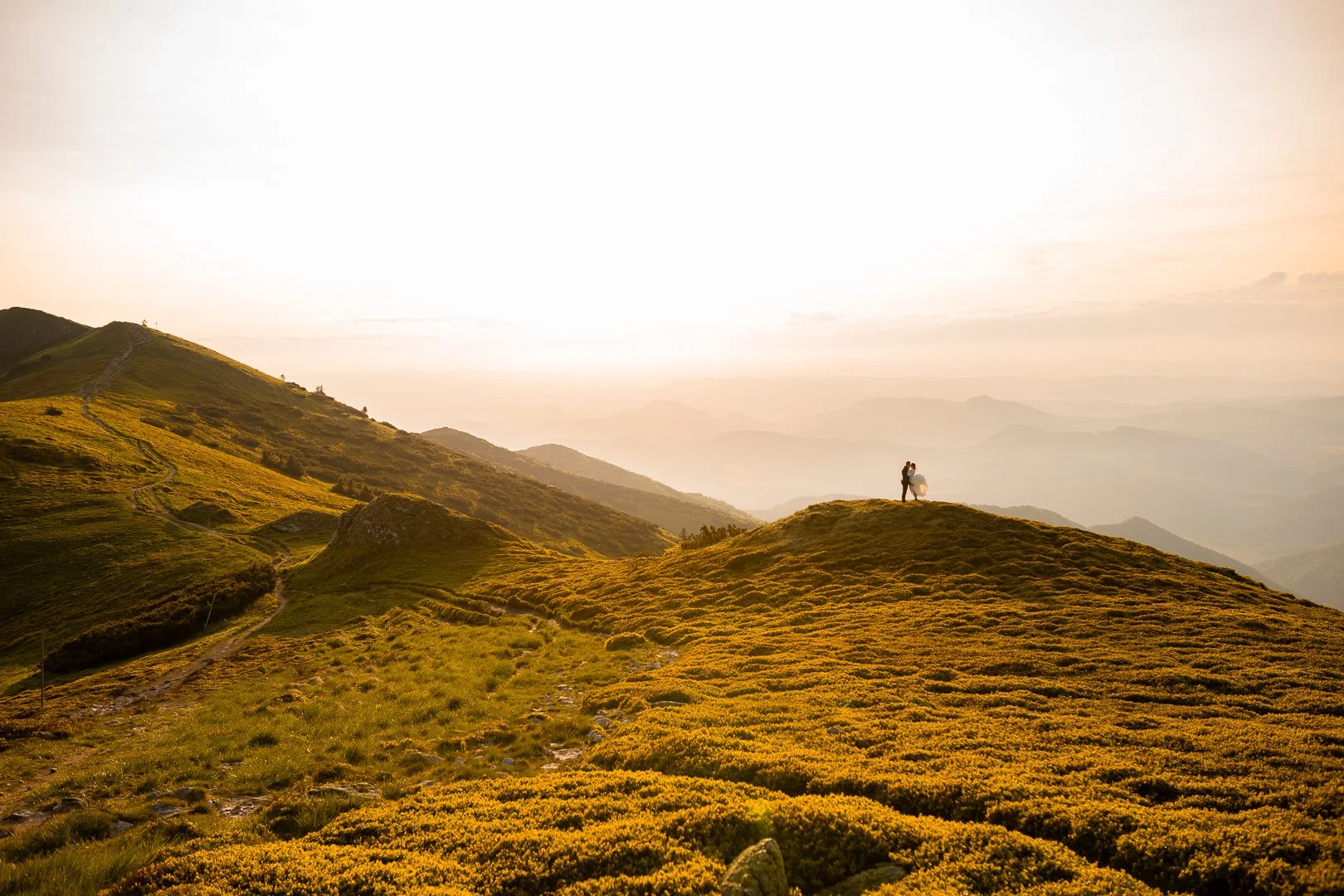 Para stoi na szczycie wzgórza, patrząc na łagodne, zielone góry podczas zachodu słońca.