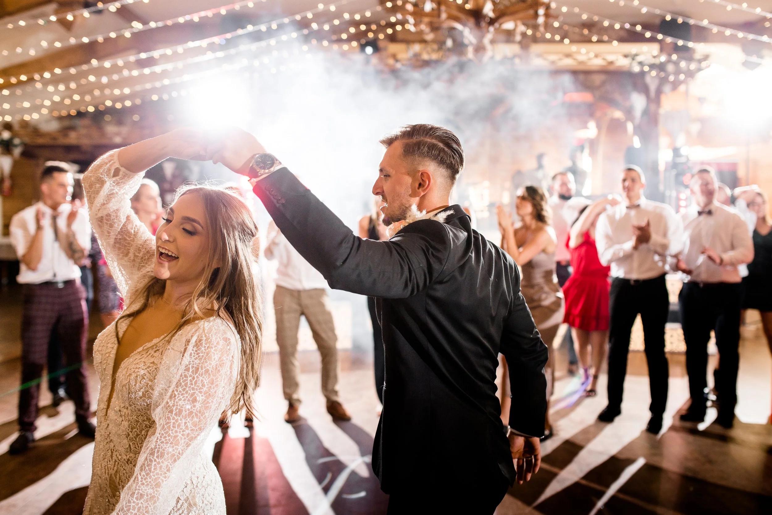 A couple is dancing happily at a wedding reception, surrounded by guests clapping and smiling under warm string lights and fog effects.