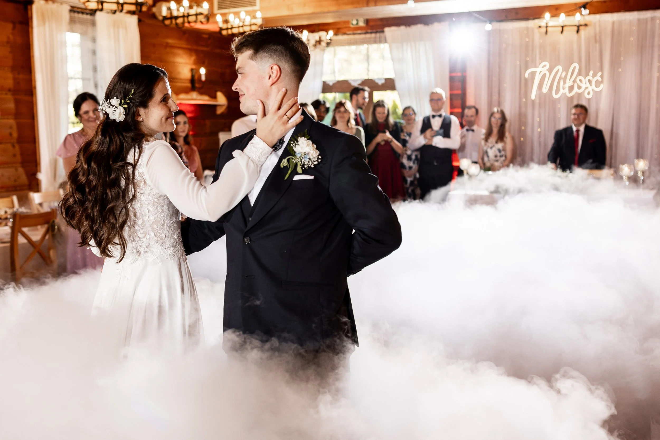 Para młoda tańczy na przyjęciu weselnym, goście obserwują taniec na udekorowanym drewnianym tle z napisem „Milosc”.