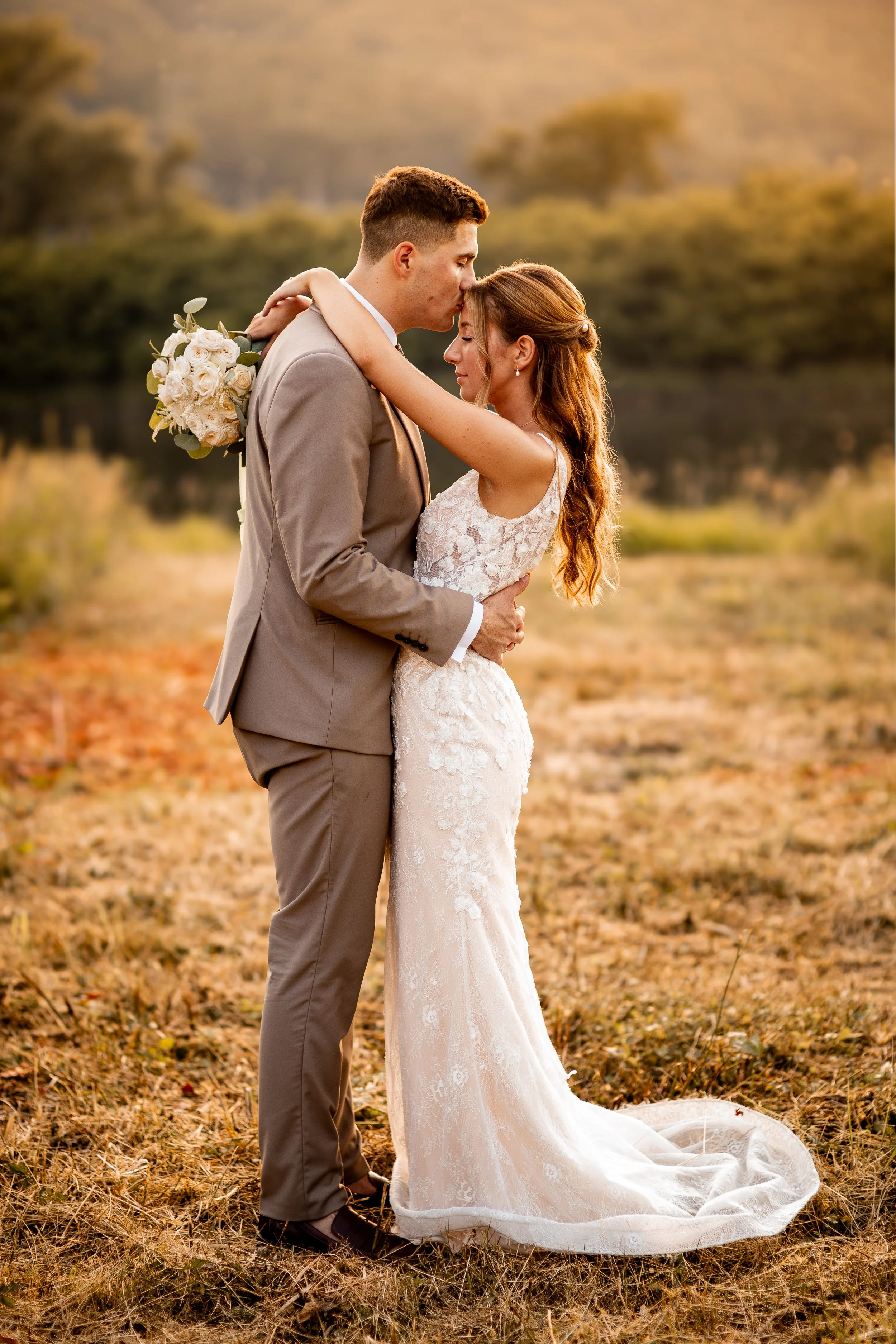 A bride and groom embrace outdoors during sunset, with the bride in a white lace wedding dress and the groom in a beige suit, holding a bouquet of white roses.