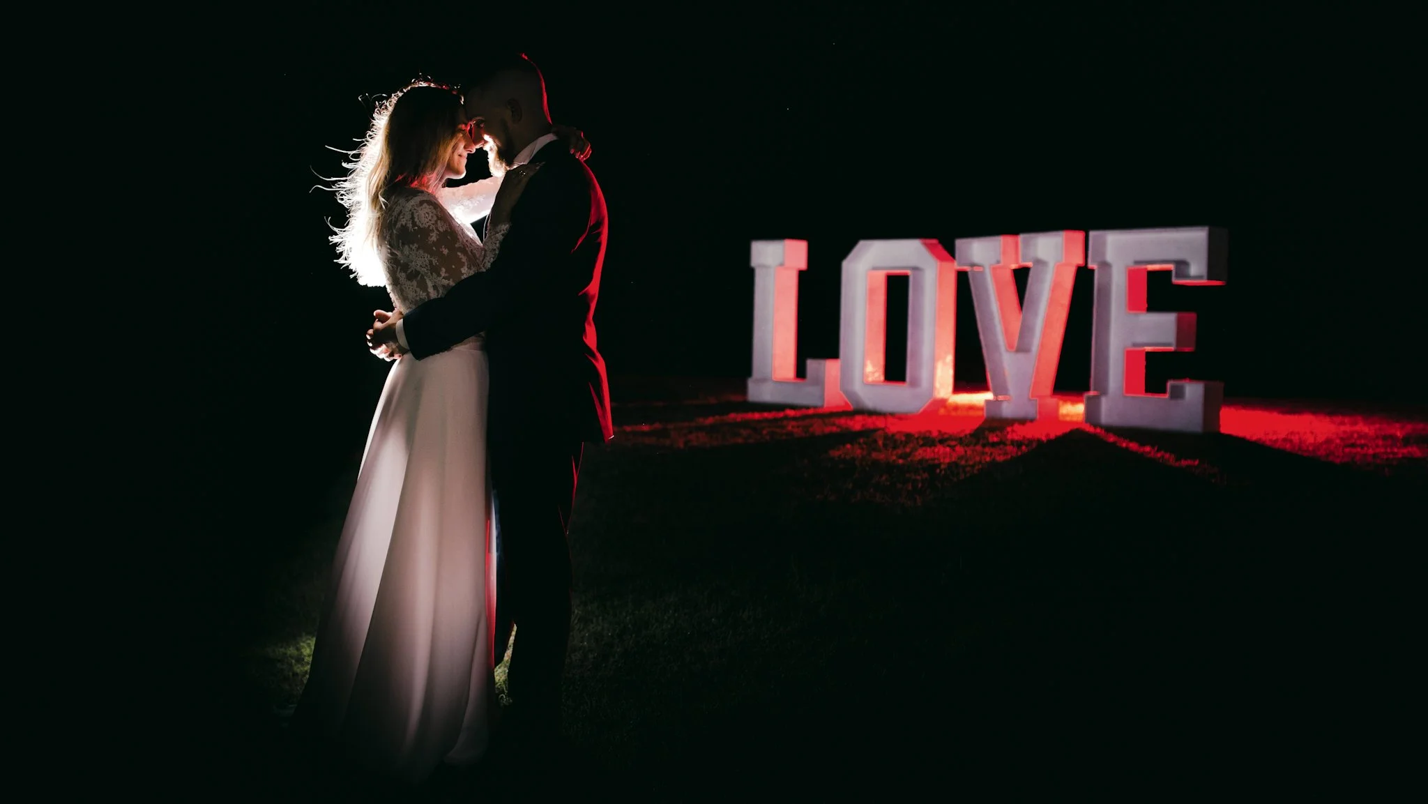 Para młoda obejmuje się na tle dużych, podświetlonych liter układających się w napis „LOVE” nocą.