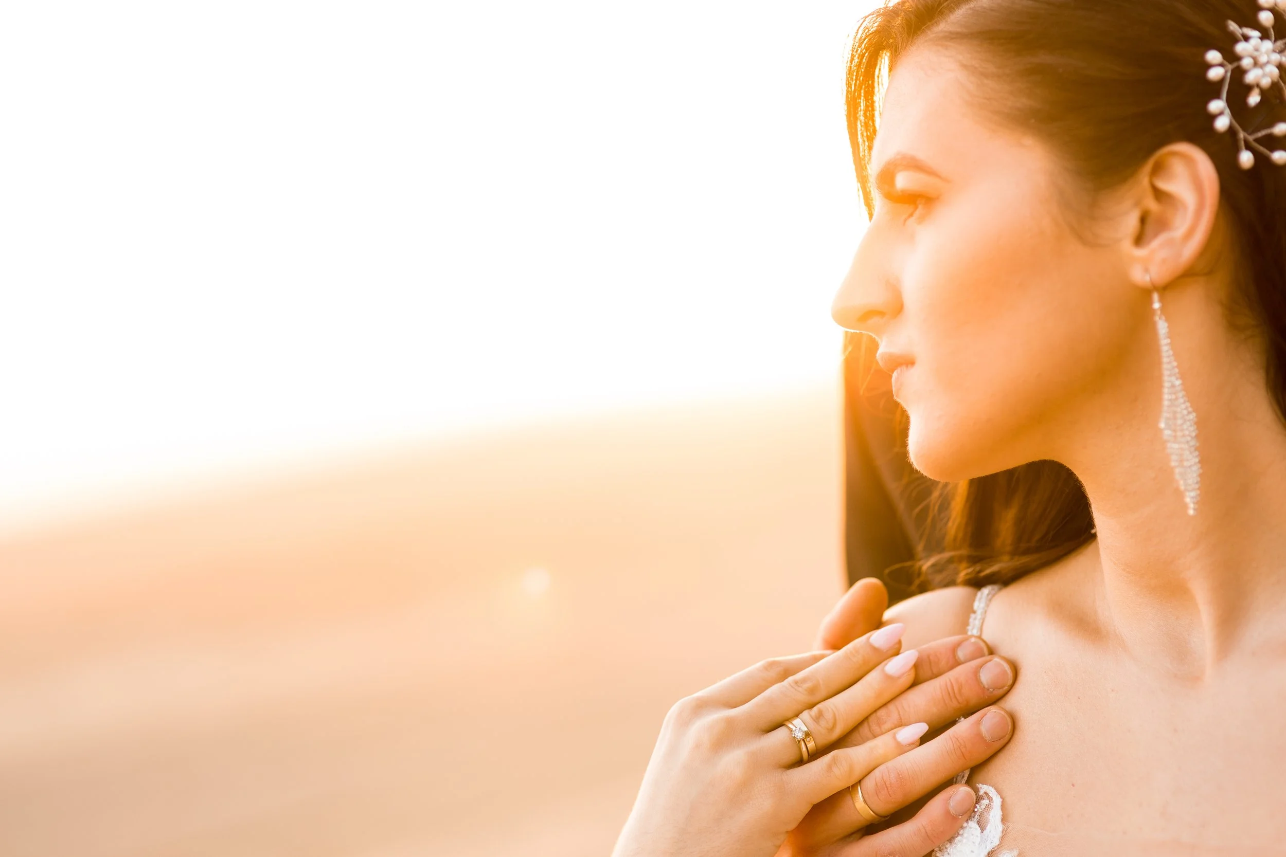 A woman with long hair and a flower hair accessory, wearing long earrings and a dress, looks to the side at sunset with her hand on her shoulder.
