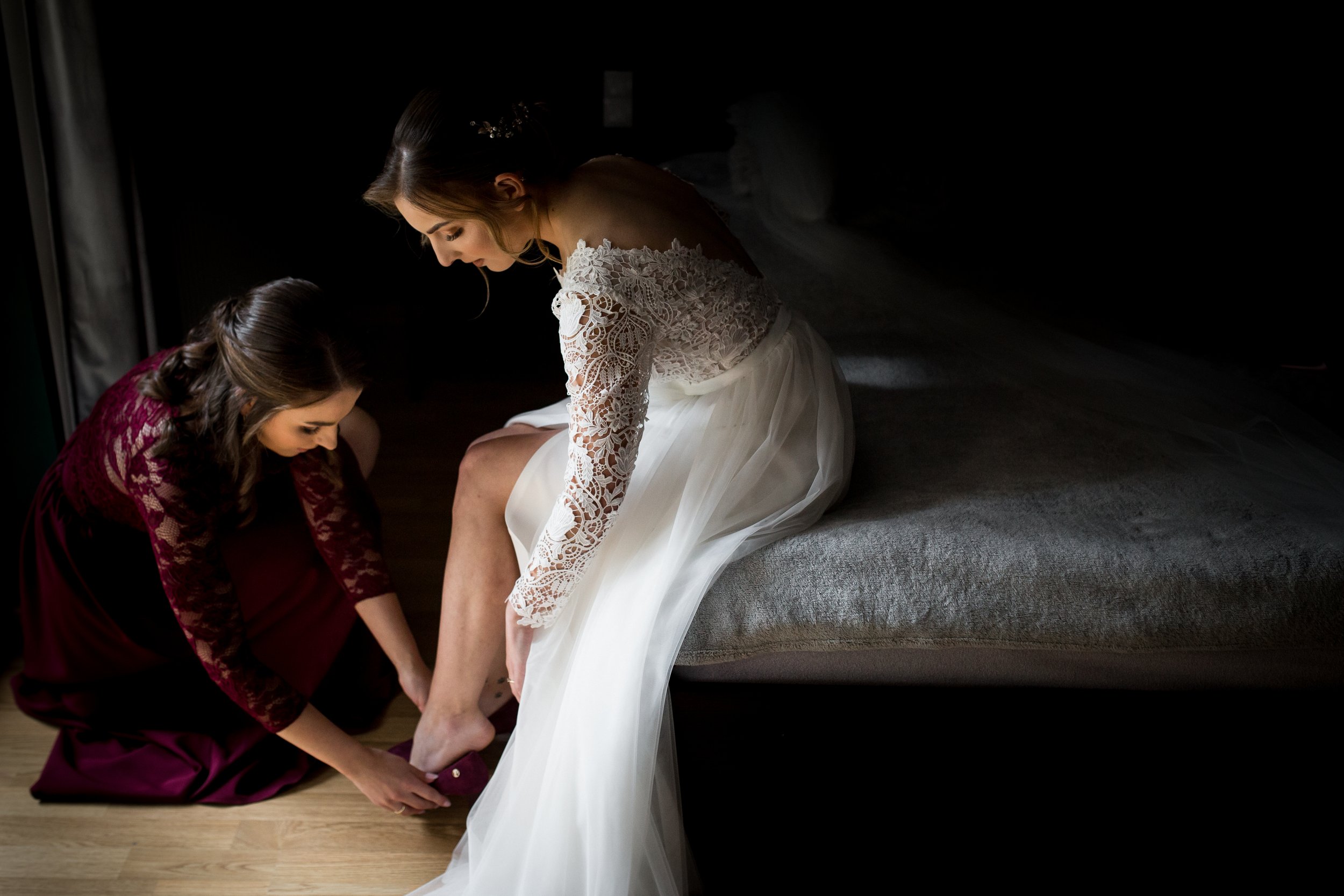 Bride in a white lace wedding gown sitting on a bed while another woman helps her with her shoes in a dimly lit room.