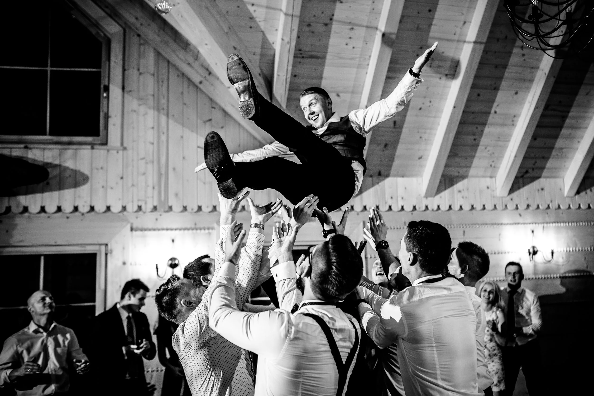 A man in formal attire being lifted by a group of people at a celebration or party in a wooden hall. The man is smiling with arms outstretched as he is being thrown into the air.